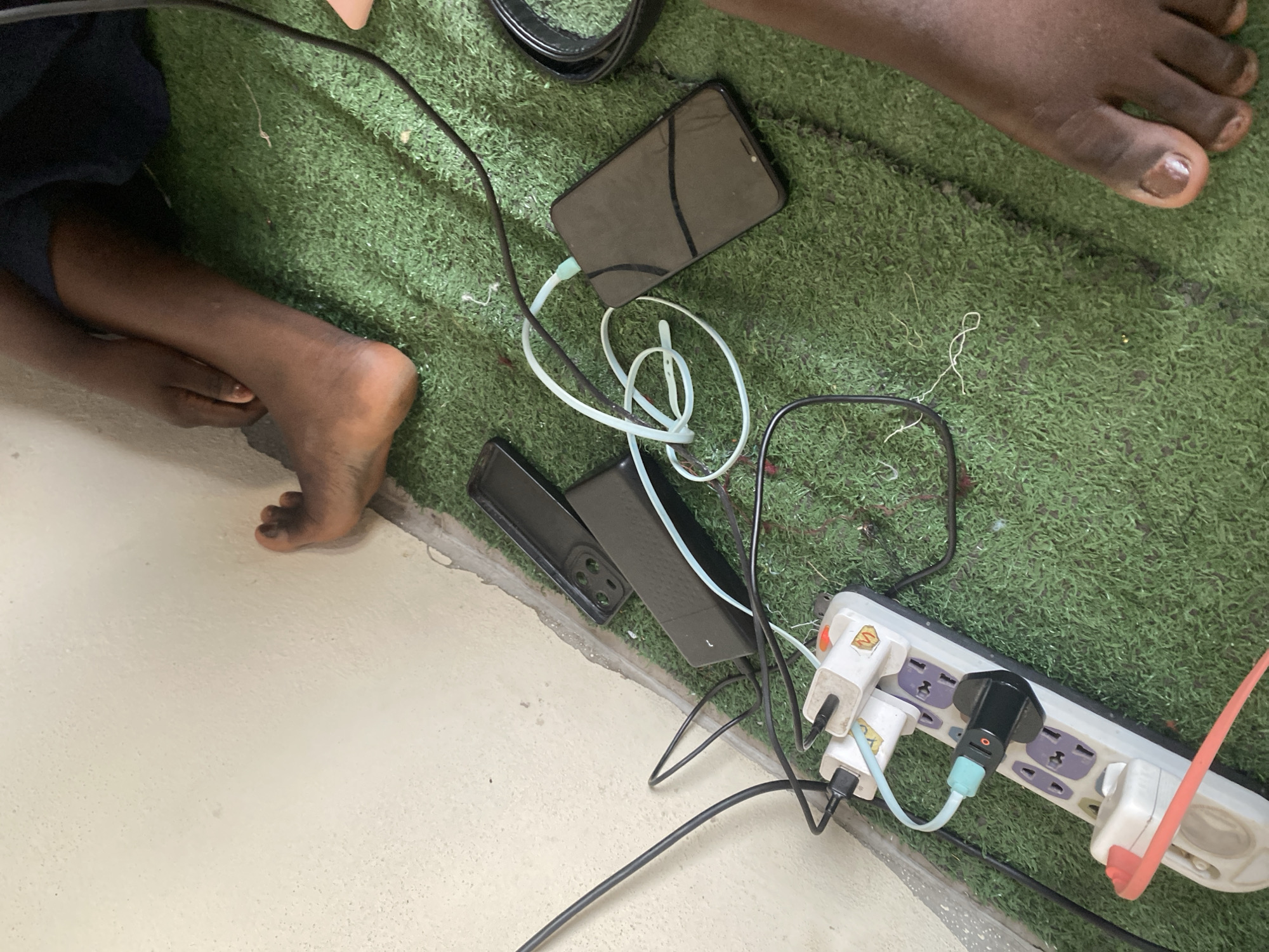 A high-angle shot shows a power strip plugged into an outlet on a wall, situated on a green, artificial turf-covered floor, presumably in Jauro Yinu, Nigeria. A person's bare feet are visible on the grass, one positioned near the wall and the other further away towards the right side of the frame. Several electronic devices and accessories are connected to the power strip, including two white power adapters with black USB ports, one black power adapter with a red indicator light, and a black smartphone charging with a light blue cable. A black smartphone case is also present on the turf. The overall scene depicts a functional charging station on a floor.