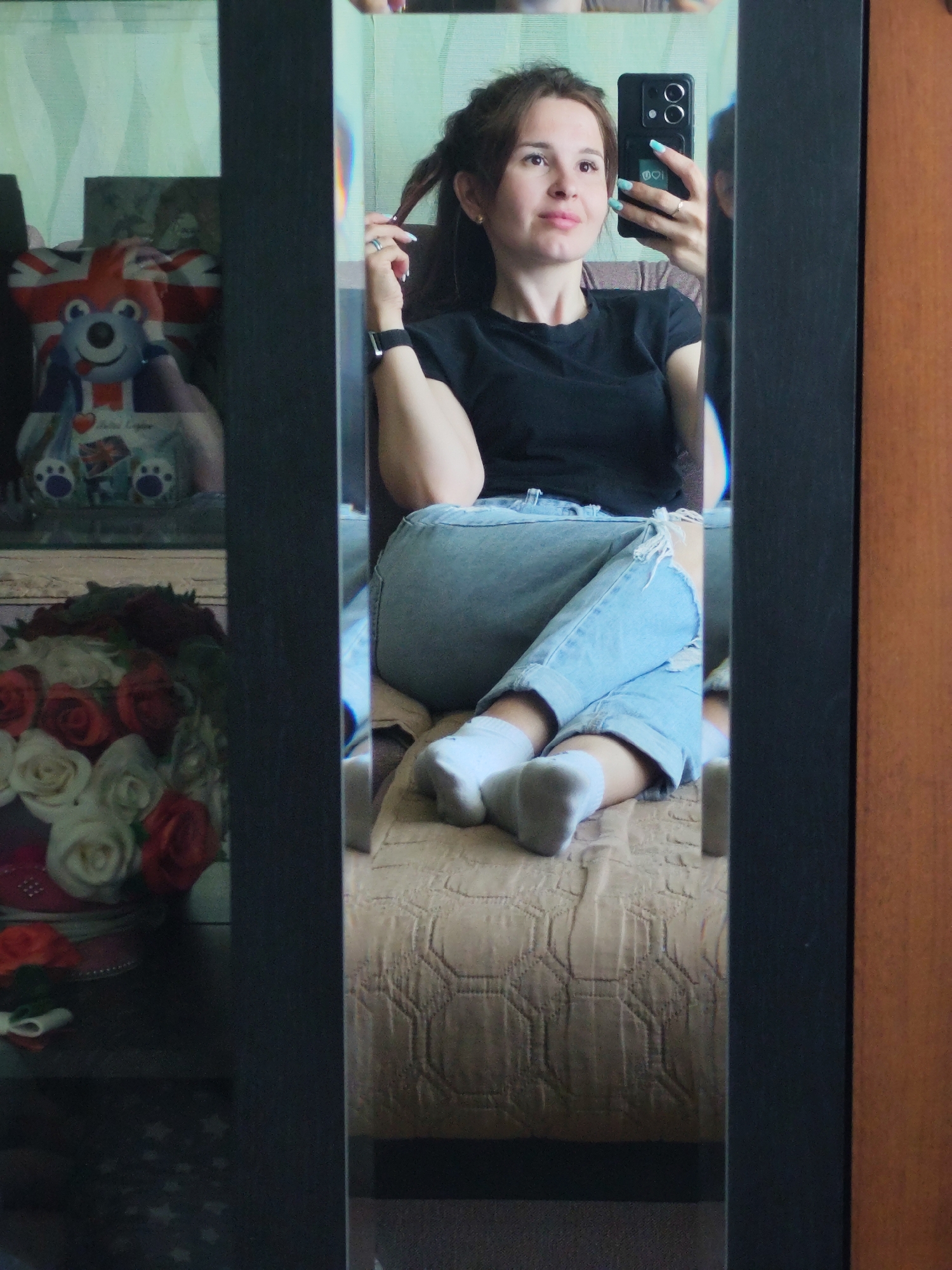A woman is taking a selfie in a mirror. She is sitting on a beige patterned surface, wearing a black t-shirt, ripped light blue jeans, and white socks. Her hair is up, and she is holding a black smartphone in her right hand. In the reflection on the left side of the mirror, there's a shelf with a teddy bear with a British flag design and a bouquet of red and white roses. A brown wooden door is visible on the right side of the mirror. The location is unidentified beyond the information provided.