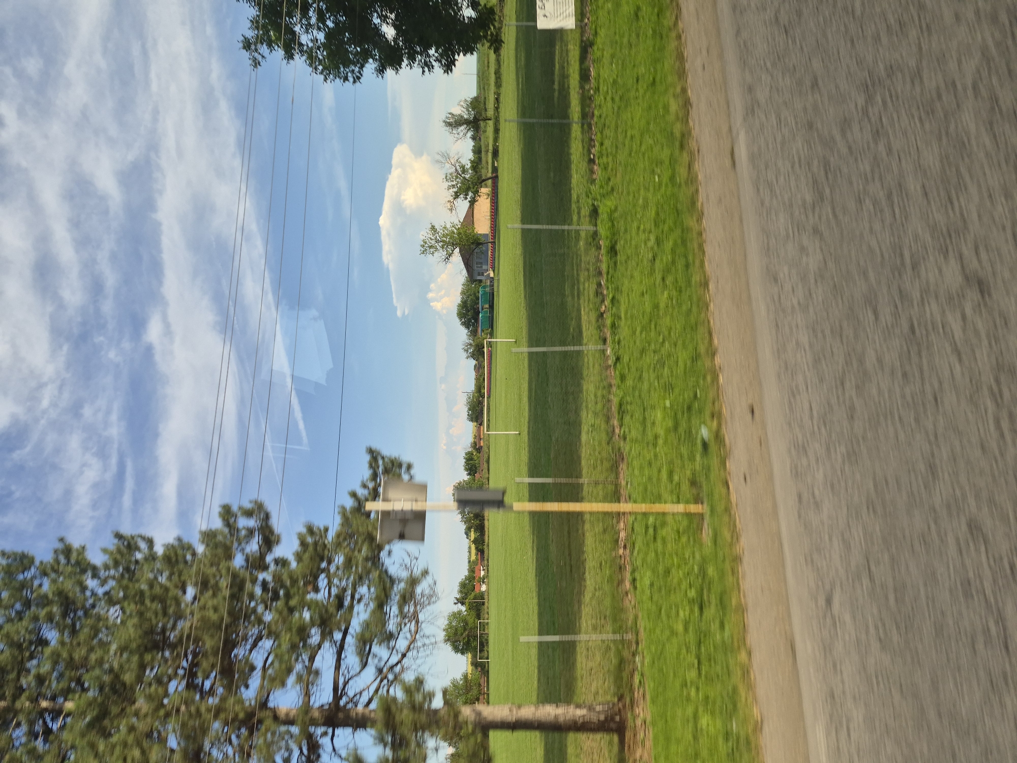 The image captures a sunny day in Obnova, Bulgaria, with a view of a well-maintained green sports field, likely a football pitch, surrounded by a fence. Several trees are visible, including a tall pine tree on the left, and a cluster of trees and buildings in the background. The sky is a bright blue with wispy white clouds, suggesting fair weather. Power lines are strung across the upper portion of the frame. The foreground is dominated by a blurred asphalt road, indicating the image was taken from a moving vehicle. There are no people or animals visible in the frame. The overall scene suggests a peaceful rural or semi-rural setting within the town of Obnova.