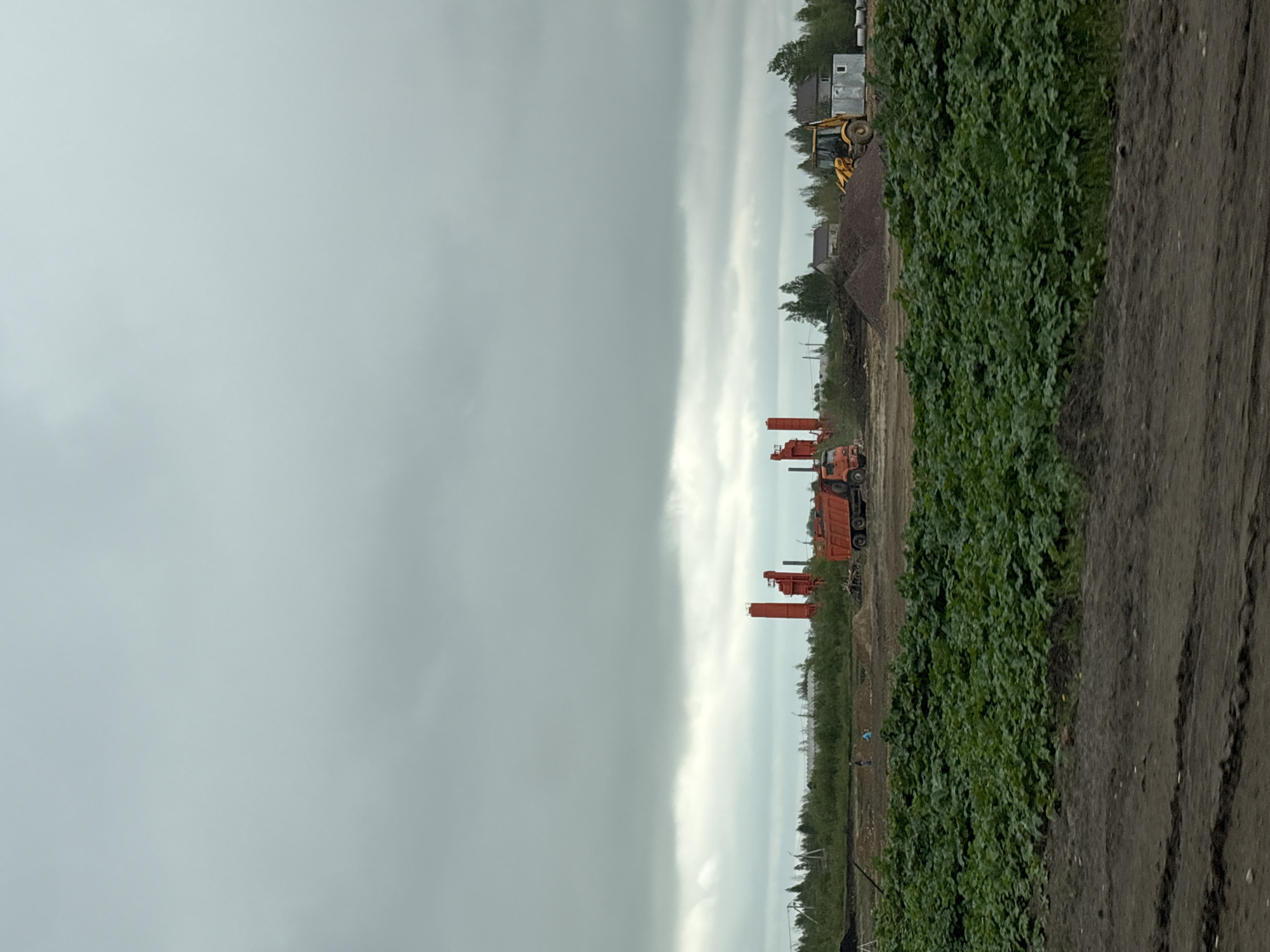 The image depicts a construction site near Myaglovo, Russia under an overcast sky. Heavy machinery including orange drilling equipment and a yellow bulldozer are present. The ground shows evidence of recent earthmoving activities. Vegetation grows along the edge of the construction area, bordering a dirt road or path. Two indistinct figures appear to be walking along the construction area. Buildings are visible in the background.