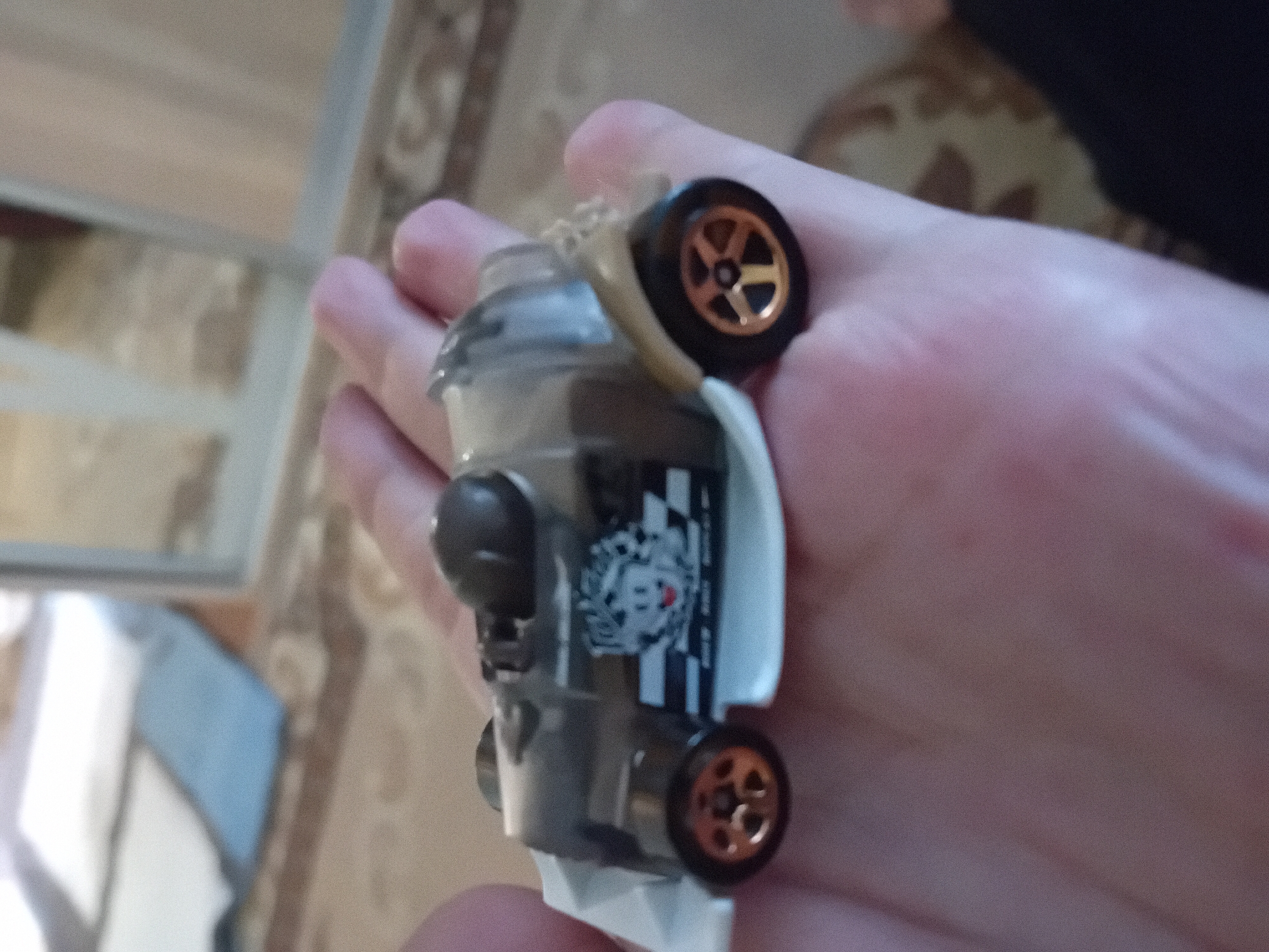 A hand is holding a miniature toy race car. The car is predominantly silver and grey with a transparent canopy. It features a black and white checkered pattern on its side with a cartoon character resembling Mickey Mouse. The wheels have gold rims. The background is out of focus and appears to be an indoor setting with a patterned carpet and a blurred reflection of a room. There is no discernible information about the city of Chuhuiv, Ukraine in the image.