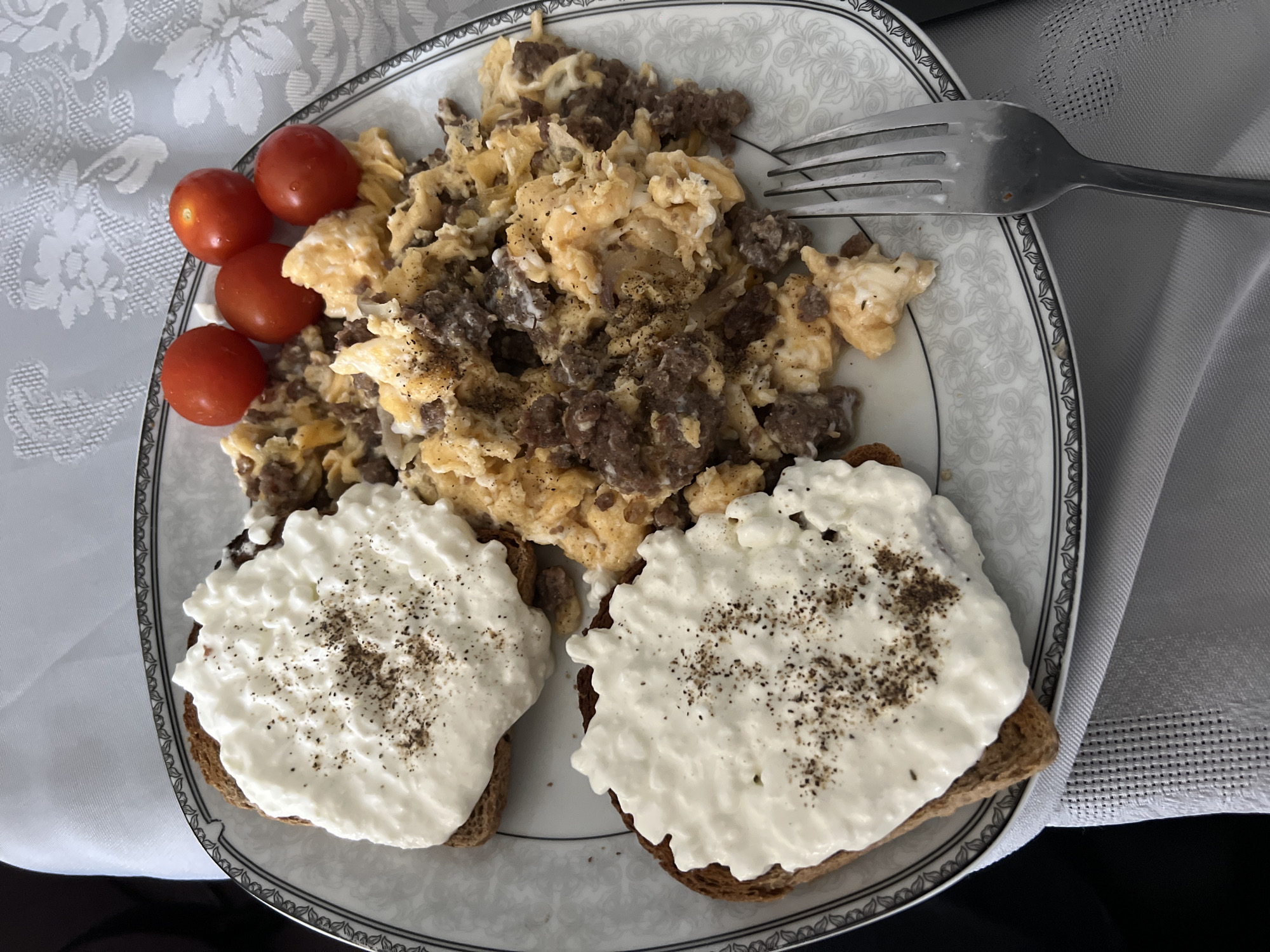 A plate of food sits on a patterned surface. The meal consists of scrambled eggs mixed with what appears to be diced beef, several small red tomatoes, and two slices of toast topped with a white, creamy substance, possibly cottage cheese, seasoned with pepper. A metal fork rests on the plate. The specific location is not discernible from the image.