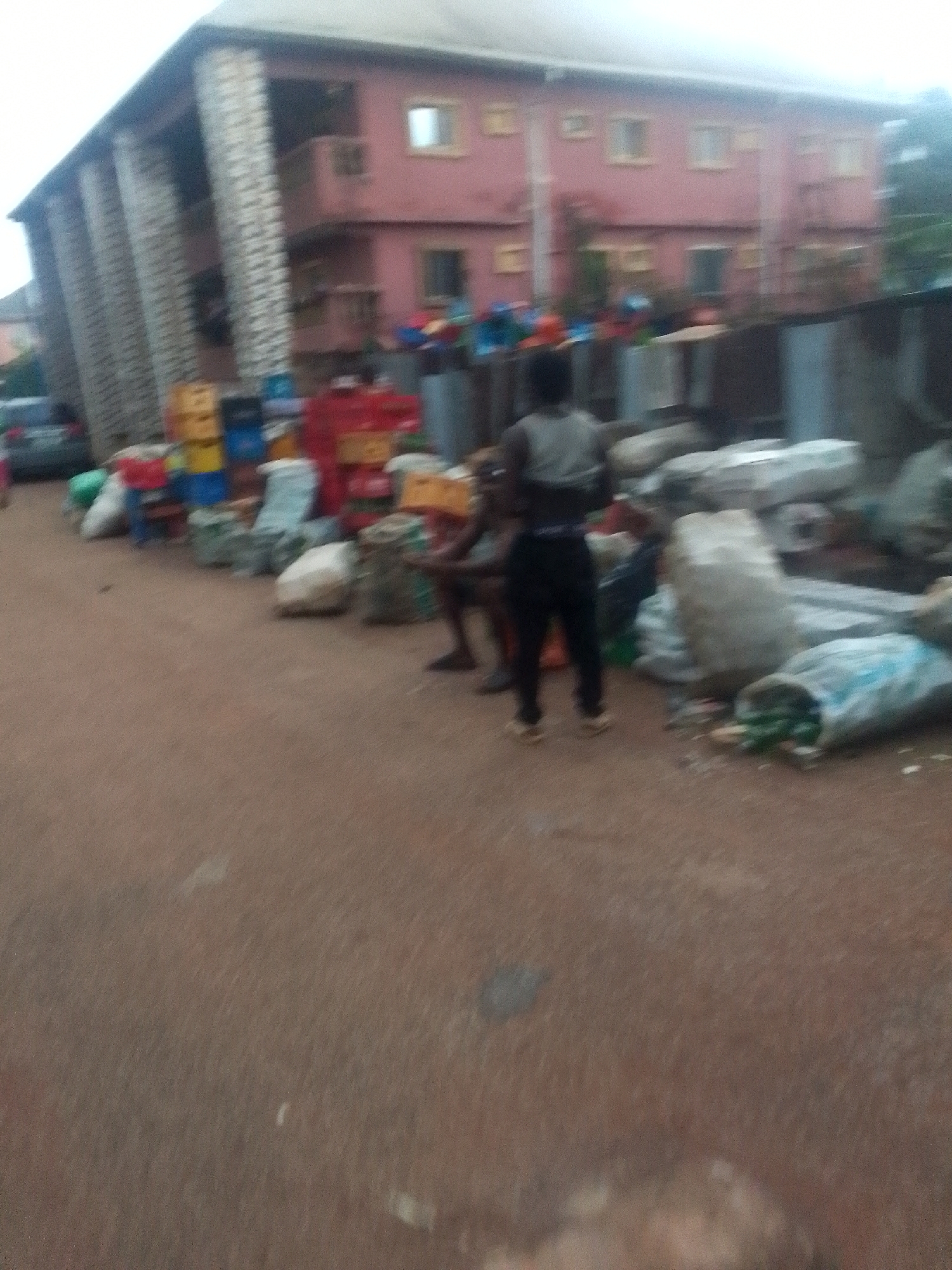 The scene depicts a street in an unspecified city and location. Two individuals are present, one standing with their back to the viewer and the other seated. A variety of objects line the street, including sacks, beverage crates, and miscellaneous trash. A two-story building with columns is visible in the background, along with a parked car. The overall impression is one of a cluttered or perhaps unsanitary urban environment.