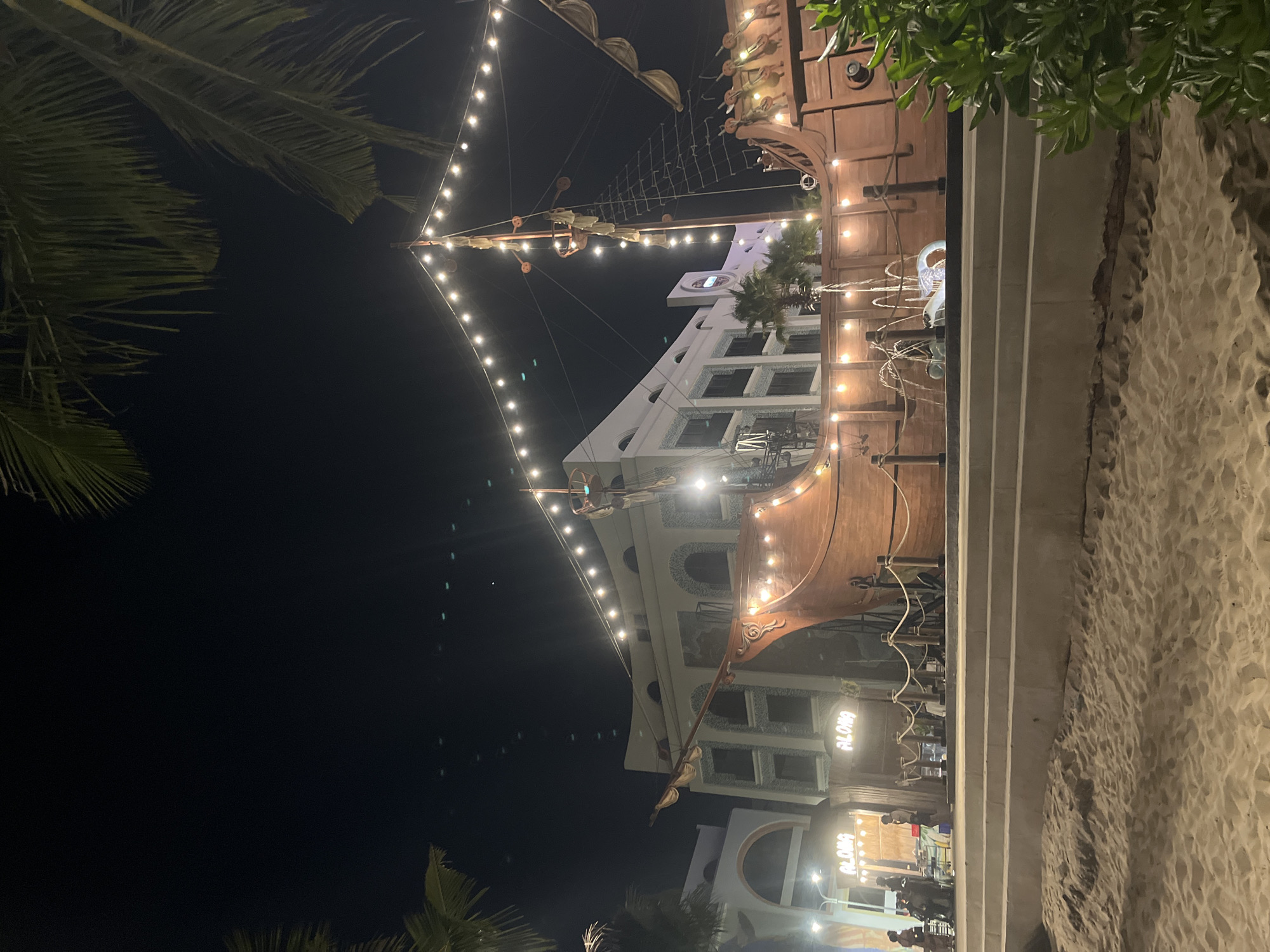 This image depicts a nighttime scene at a beachside establishment in Nha Trang, Vietnam, that appears to be designed with a pirate ship theme. In the foreground, sand is visible, with a set of concrete stairs leading up to a raised walkway. Beyond the stairs, a large, wooden pirate ship structure dominates the scene. The ship is illuminated by strings of warm white fairy lights that drape across its masts and hull. Fountains are actively spraying water in front of the ship. In the background, a modern building with arched windows and a white facade is visible, also decorated with lights. Two signs with the word "ALOHA" in illuminated lettering are mounted on structures near the building. Several people are present, some standing and observing the scene, while others appear to be seated. Palm fronds hang from the top of the image, suggesting a tropical location. The sky is dark, indicating it is nighttime.