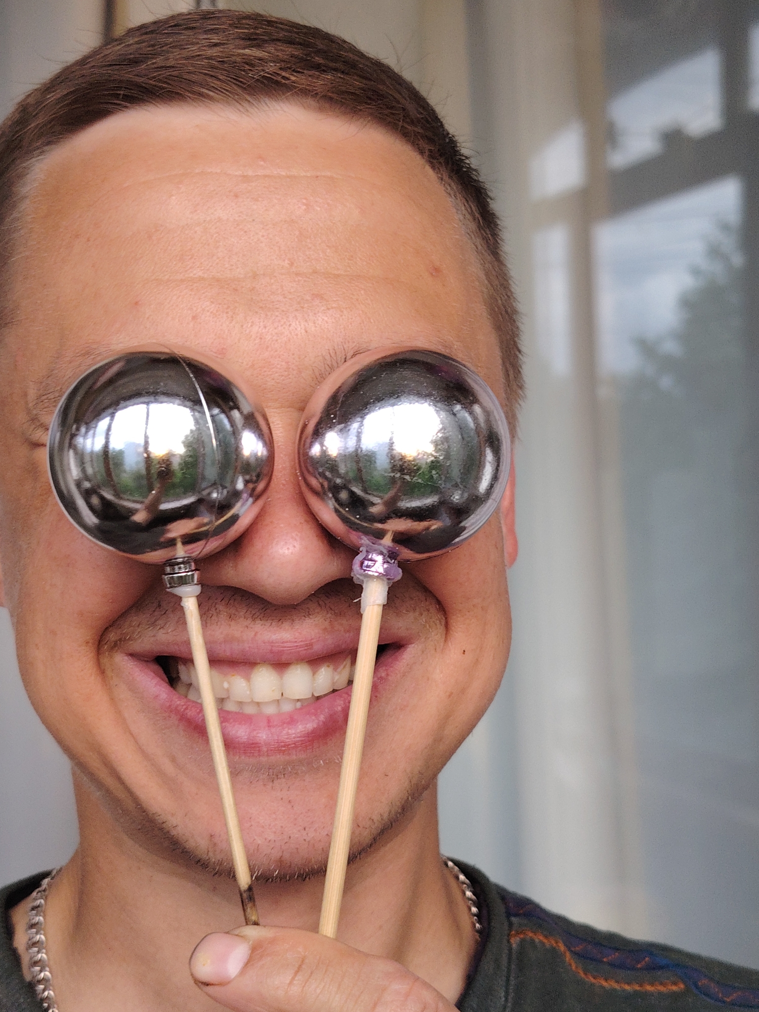 A close-up portrait captures an adult male in Chaikovsky, Russia, playfully holding two shiny, spherical, metallic objects on wooden sticks in front of his eyes, effectively obscuring them. The man is smiling widely, revealing his teeth, and holds the two sticks with his right hand, positioned beneath his chin. He has short brown hair and light stubble around his jawline.
The metallic spheres are highly reflective, prominently showing reflections of a bright outdoor scene with trees and what appears to be a window frame, along with a cloudy sky. The background behind the man is softly blurred, revealing a window through which an outdoor scene with light and possible foliage is visible, indicating it is daytime. The man is wearing a dark t-shirt with patterned detailing on the shoulder and a silver chain necklace. His expression conveys humor and lightheartedness.