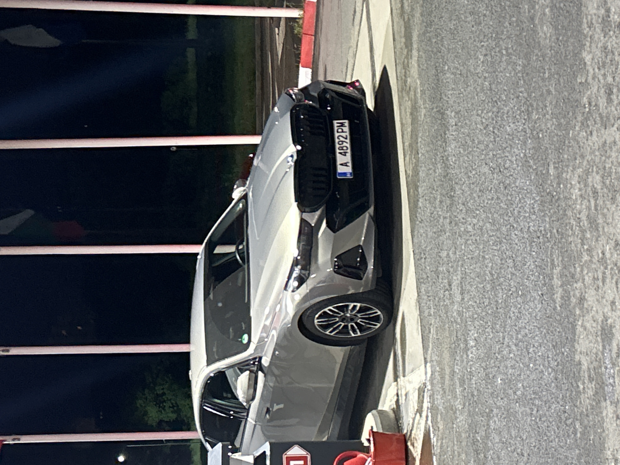 A gray BMW car is parked on a paved surface, presumably a street. The license plate reads "A 4892 PM." The car appears to be in Burgas, Bulgaria, as inferred from the vehicle registration. There are vertical white poles with red stripes visible in the background, which could be part of a structure like a car wash or a barrier. A red and white object is visible near the front wheel of the car, but is unidentifiable. The lighting suggests the photo was taken either at night or in a dimly lit area.
