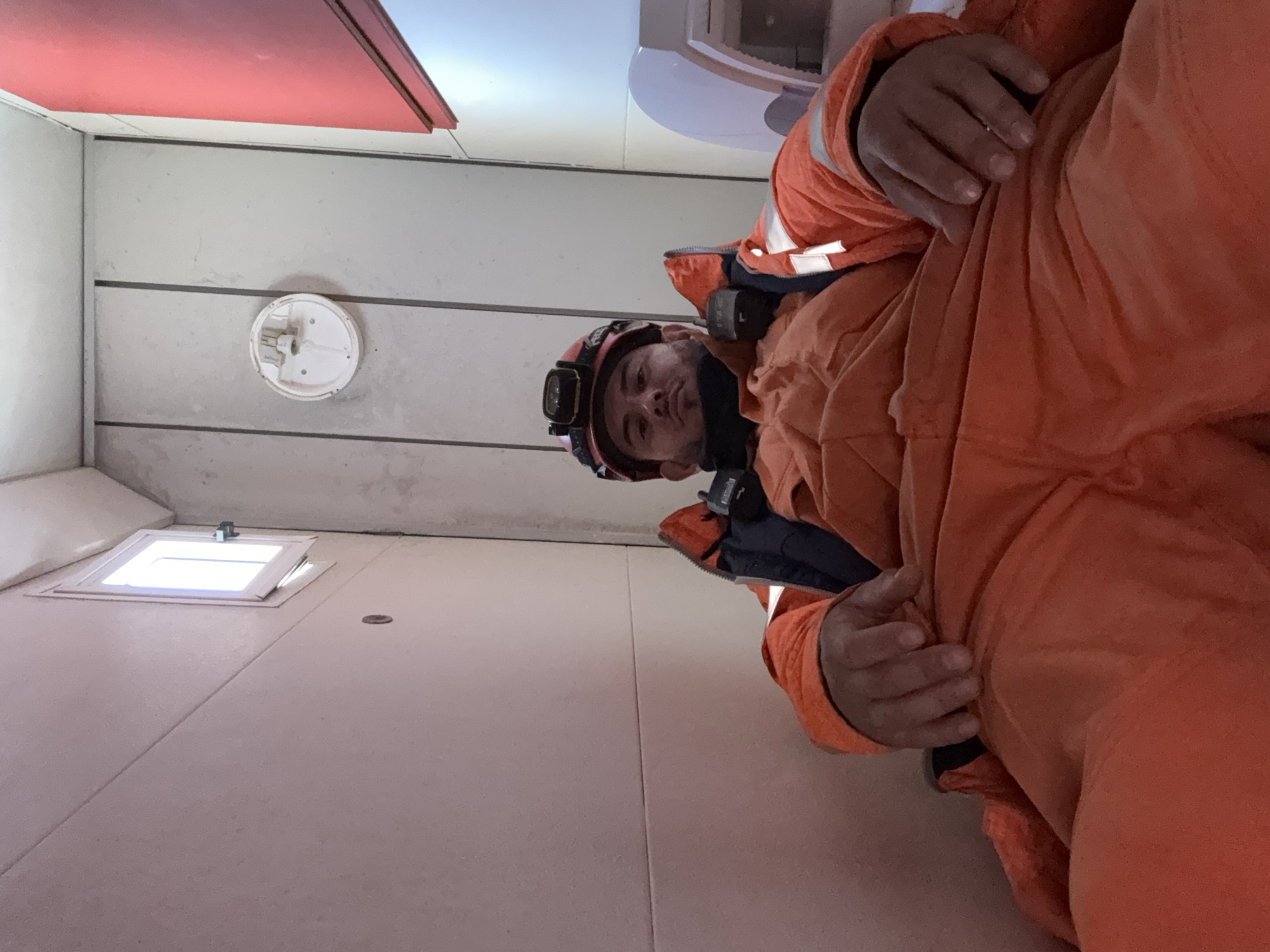 A man wearing orange overalls and a headlamp is looking down at the camera. He is in a room with white walls, a small square window, and a round white device mounted on the wall. A red object is visible on the ceiling. The location is unknown and there is no information about Brest, France.