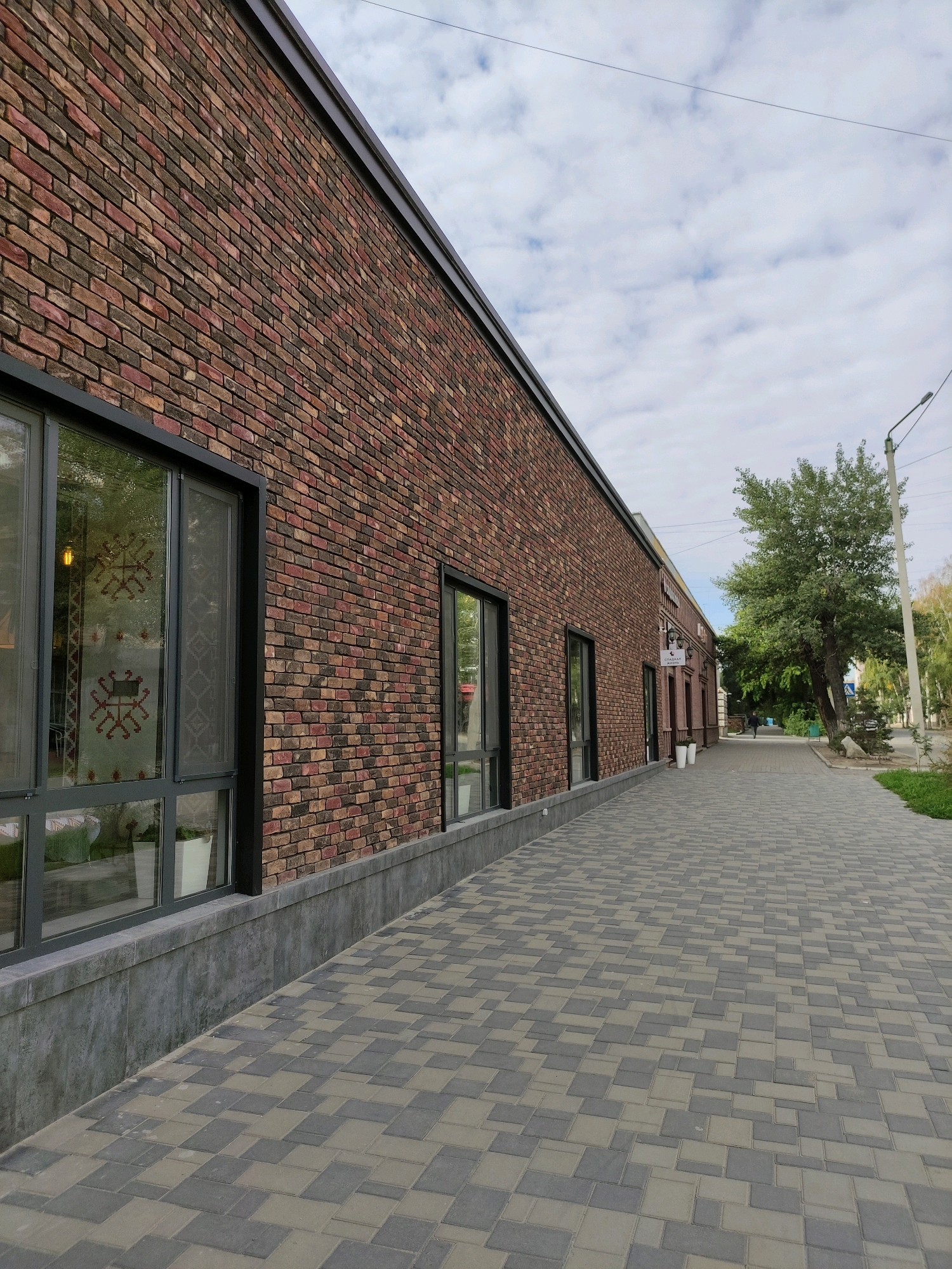 The image depicts a modern building with a brick facade, large windows, and a paved sidewalk in front of it. The building appears to be commercial, possibly a cafe or restaurant, as suggested by a sign visible on the right side of the facade with Cyrillic lettering. The sidewalk is constructed with interlocking concrete pavers in a geometric pattern of light and dark gray. A street lamp stands on the right side of the sidewalk, and trees line the street in the background. The sky is partly cloudy. The scene is set in the city of Semey, Kazakhstan, at a location that could be along a pedestrianized street or commercial area. There is one person walking away from the camera on the sidewalk in the distance. No other individuals or specific actions are clearly discernible.