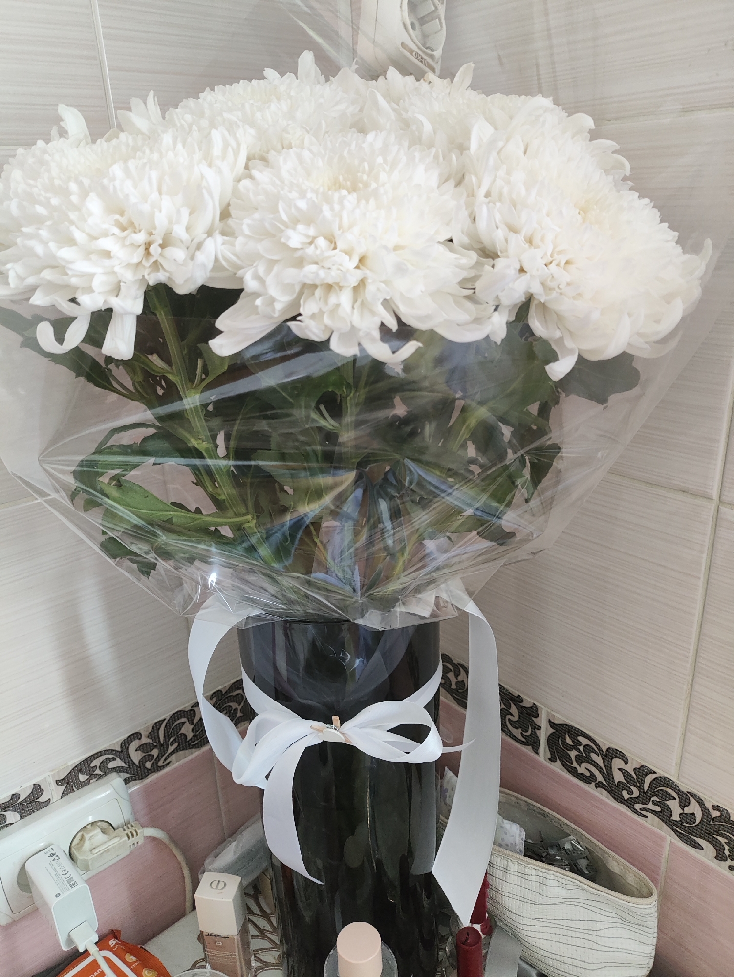 In a tiled room, a bouquet of white chrysanthemums, wrapped in clear plastic and tied with a white ribbon, stands in a dark vase. To the left, an electrical outlet with a white adapter is visible, along with a charger and a small orange package. A cosmetic box and a beige woven bag are situated to the right of the vase. The background consists of pale pink and white tiles. The scene appears to be an interior setting, possibly a home or hotel room, in Kharkiv, Ukraine.
