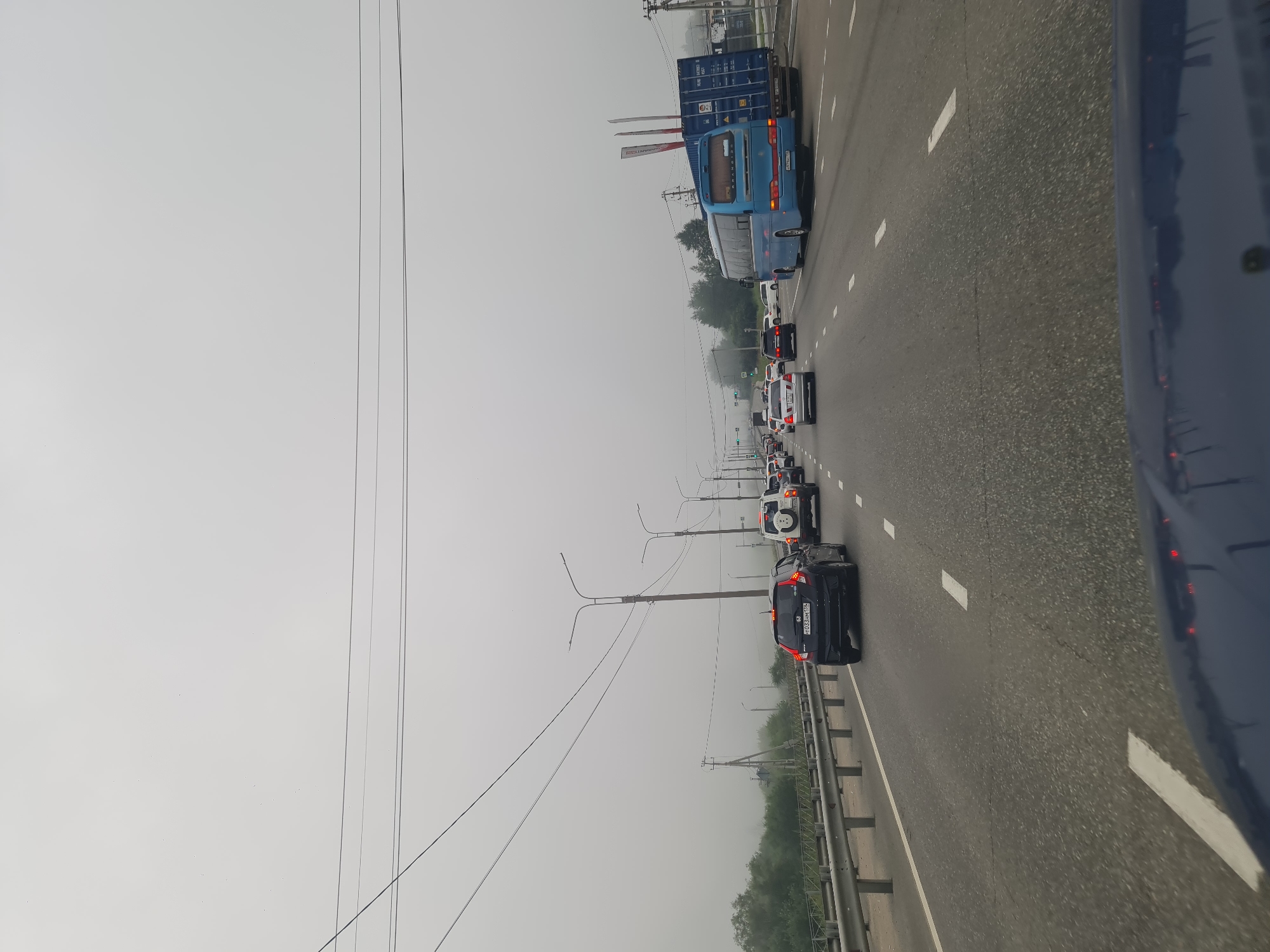 The image depicts a view from a vehicle driving on a multi-lane highway in a foggy or overcast condition. Several cars are visible on the road, including a black Honda Fit in the foreground, a white SUV, and a blue bus towing a shipping container. The highway is bordered by a metal guardrail on the left, with trees and vegetation beyond it. Utility poles and power lines are also present. The scene appears to be on the outskirts of Irkutsk, Russia, given the style of license plates and the general landscape. There are no people clearly visible, and the overall atmosphere is one of normal traffic flow under challenging visibility conditions.