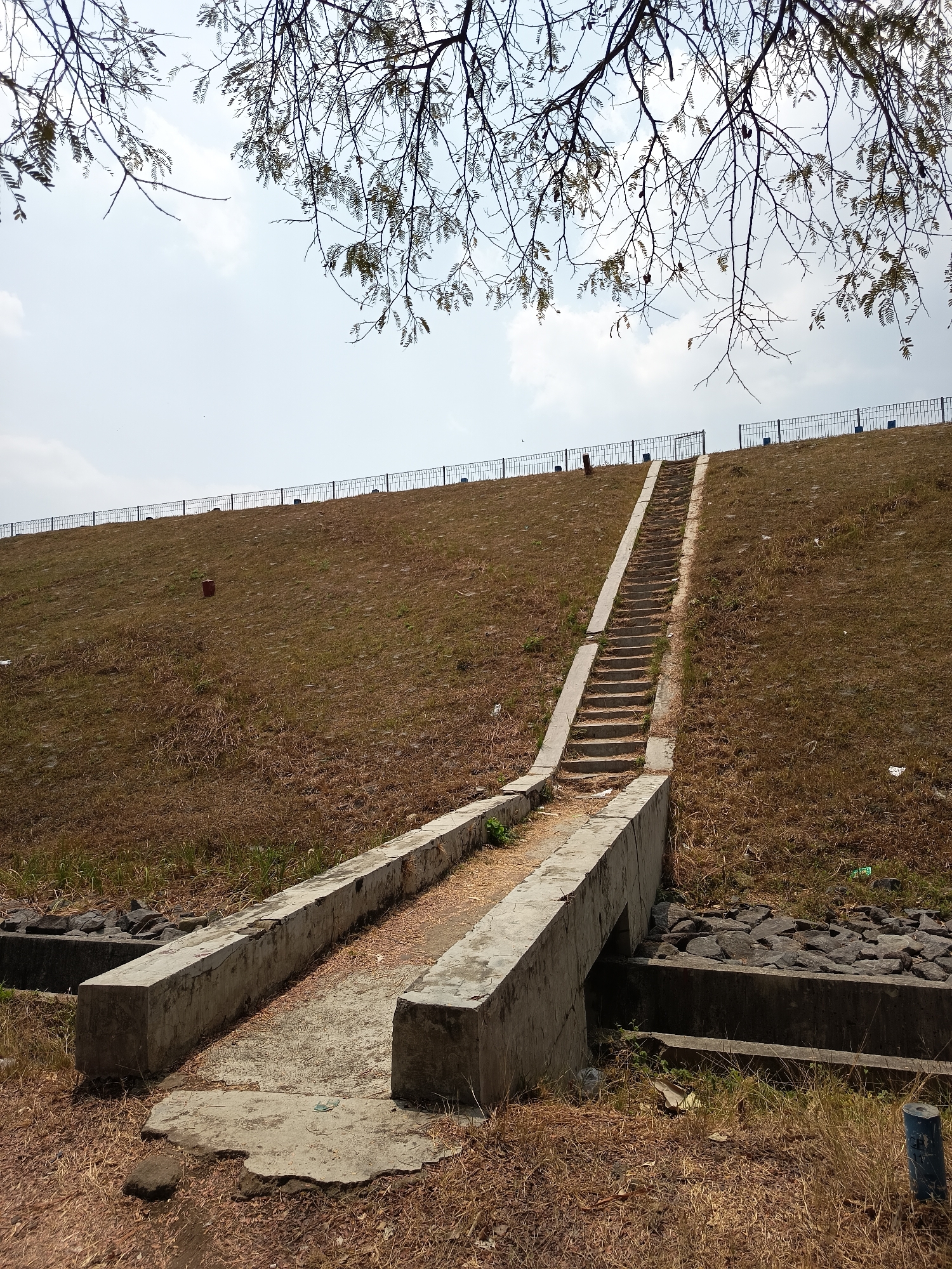 A wide-angle, low-angle shot captures an outdoor scene featuring a concrete staircase ascending a grassy embankment in Lalung, Indonesia. The embankment is covered in dry, brown grass with patches of green vegetation. A black metal fence lines the top of the embankment. The sky is bright and cloudy, with the bare branches of a tree extending into the frame from the top. The staircase itself is made of concrete with concrete railings on either side. At the base of the staircase, there are two parallel concrete structures that appear to form a walkway or channel. Rocks are visible in a concrete channel to the right of the walkway. Debris is scattered across the ground and on the embankment. The scene appears to be a public park or recreational area.