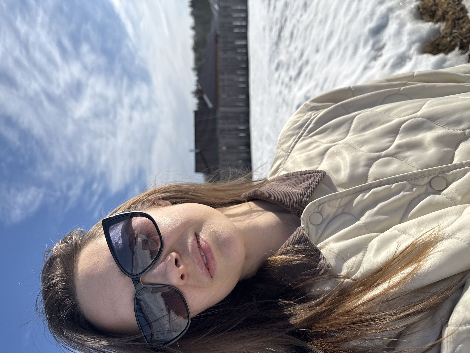 A female subject is depicted in a close-up selfie taken outdoors in Chaikovsky, Russia, during daylight hours. She has long, straight brown hair, wears black cat-eye sunglasses, and her lower face shows a neutral expression with slightly parted lips. Her attire includes a light beige quilted jacket with a brown corduroy collar and white buttons. Reflections in the sunglasses show a cloudy sky and the camera device. The background features a ground partially covered in melting snow, revealing patches of brown earth. Behind this, a dark wooden fence runs horizontally, beyond which several residential houses with dark roofs and a line of trees are visible. The sky is bright blue with scattered white, wispy clouds.