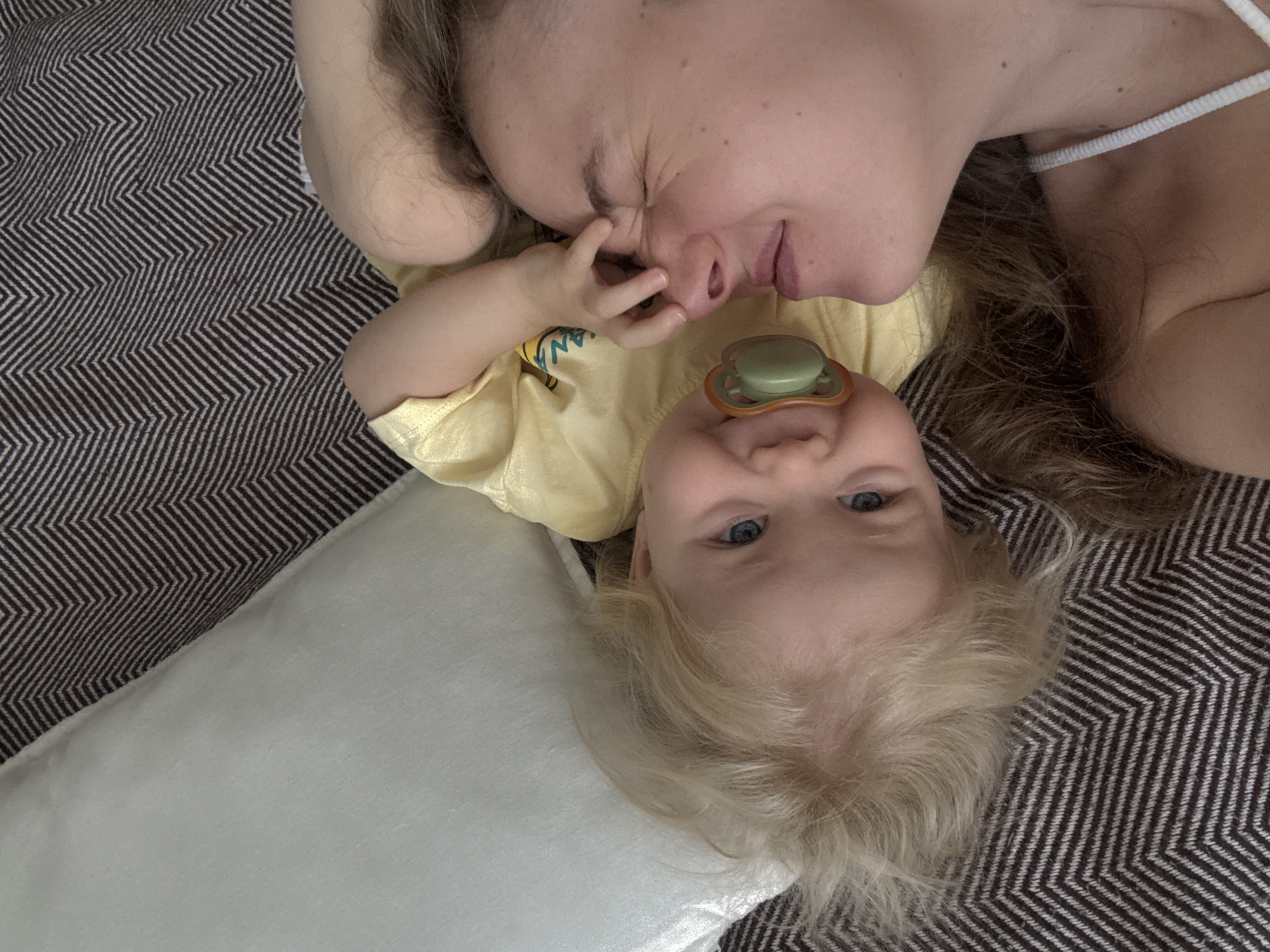 A woman and a toddler lie on a textured blanket, the woman's face turned towards the toddler. The toddler, wearing a yellow shirt and a pacifier, looks at the woman, with one hand touching her nose. A white pillow is visible to the left. The scene appears to be indoors, possibly a bedroom. The image is taken from a high angle, looking down at the subjects. There is no information in the image that would indicate a location in Vladimir, Russia.