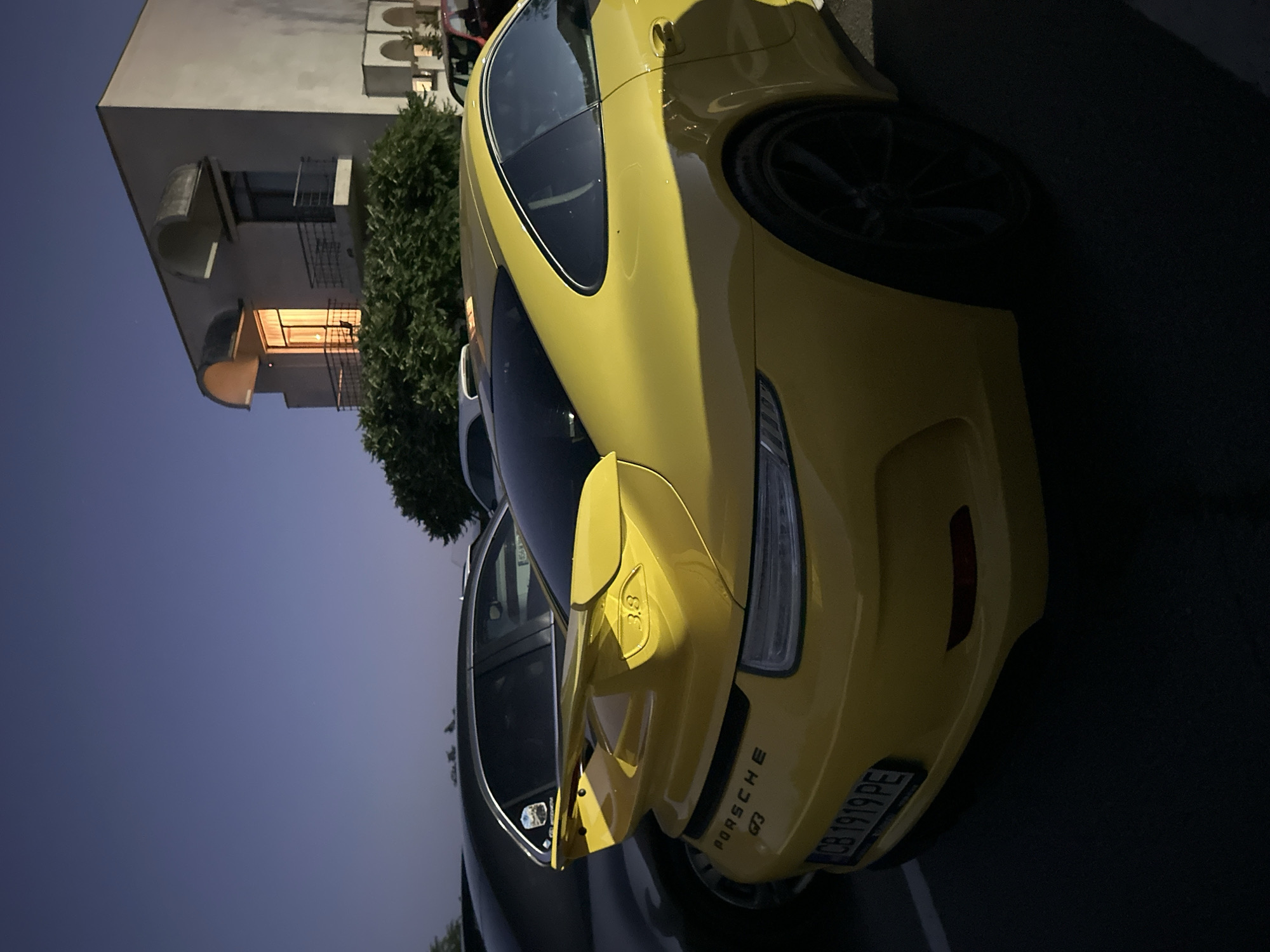 The image captures a yellow Porsche 911 GT3 parked in what appears to be a parking lot at dusk, in front of a building in Arbanassi, Bulgaria. The car is positioned at an angle, showing its rear passenger side. The building is a multi-story structure with a light-colored facade, featuring arched window openings and balconies. The sky is a deep blue, indicating the transition from daylight to night. There are other vehicles visible in the background, partially obscured by trees and shadows. The license plate of the Porsche is clearly visible and reads "CB 1919 PE". The overall scene is dimly lit, with artificial lights emanating from within the building.