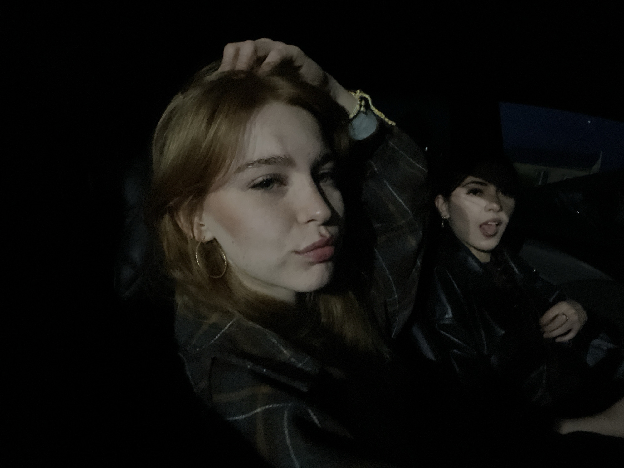 In the original image, two individuals are captured in a dimly lit environment, possibly inside a vehicle. The person in the foreground has reddish-brown hair and is looking towards the camera, with their hand resting on their head. They are wearing a plaid shirt. To the right and slightly behind is another individual wearing a black leather jacket and a dark hat. Their tongue is sticking out, and their gaze is directed towards the camera. The background is predominantly dark, with faint hints of outdoor lighting or streetlights visible in the upper right portion of the frame, suggesting an urban nighttime setting. However, there are no discernible landmarks or clues within the image that would allow for the precise identification of the location within the city of Пруды, Russia.