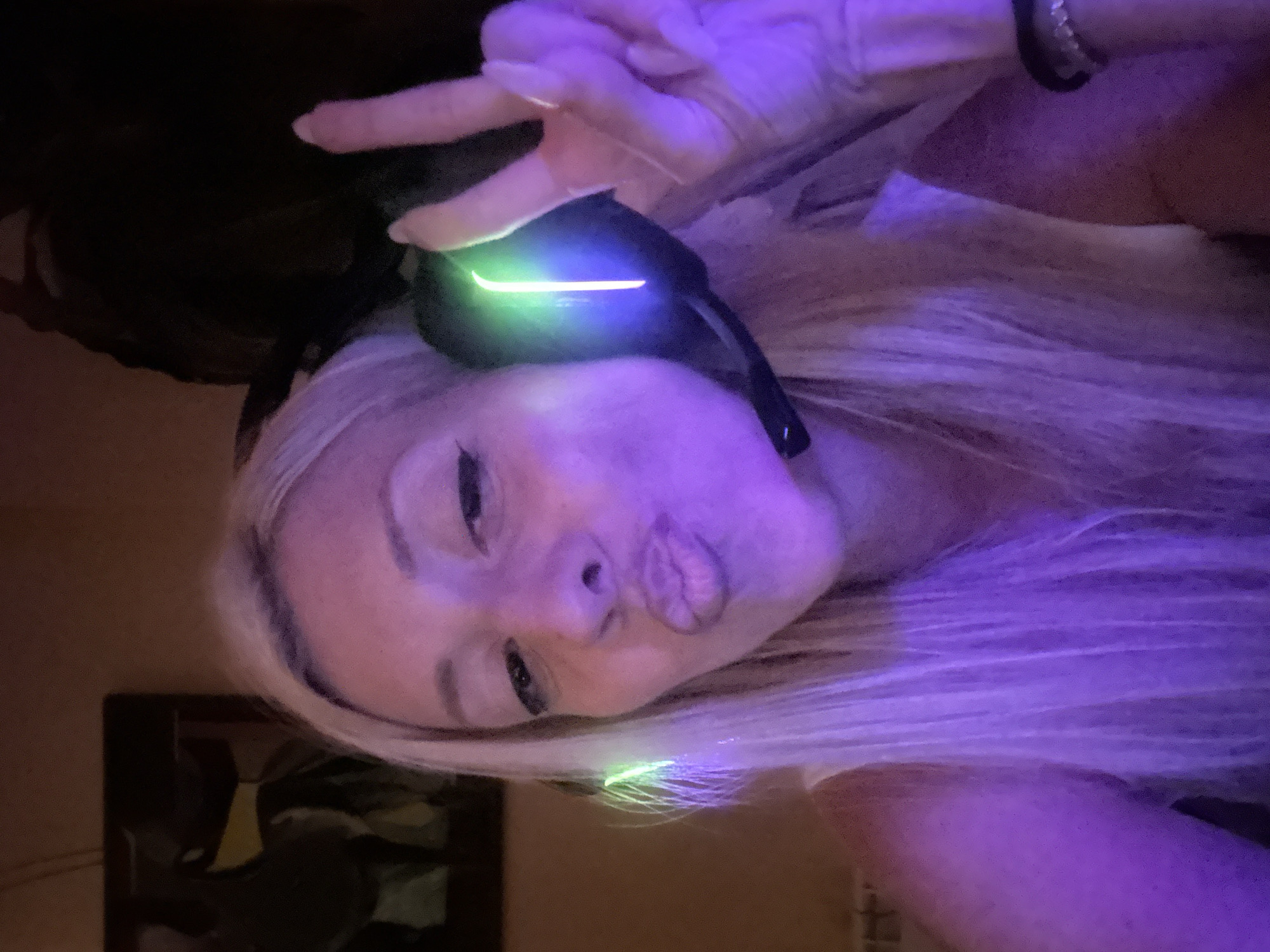 The image is a close-up, low-angle selfie of a woman with blonde hair wearing headphones. She is puckering her lips and making a peace sign with her right hand. The headphones have glowing green accents. The lighting is predominantly purple and green, suggesting a low-light environment possibly with ambient lighting from the headphones or other sources. There is a hint of a painting on the wall in the background, on the left side of the image. The woman is wearing a white top. The overall impression is a casual, playful self-portrait. The location in the city of Pazardzhik, Bulgaria cannot be determined from the image itself.