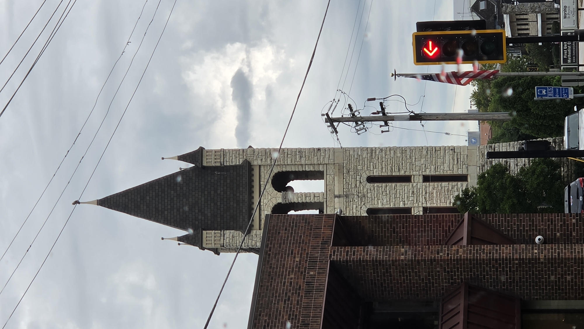 The image shows a street view in Waukesha, Wisconsin, USA, on a cloudy day. In the foreground on the left is a brick building with a dark roof. To the right of the brick building is a tall, stone church tower with a dark, pointed roof and small spires. The tower has arched openings and tall, rectangular windows. Above the church tower, a bird is perched on a finial. Several power lines cross the sky. In the midground, there is a utility pole with electrical equipment and two flags, one of which is an American flag. A traffic light is visible on the right, displaying a red left arrow. Behind the utility pole and flags, trees and other buildings are visible in the background, including what appears to be the First United Methodist Church. Several vehicles are parked on the street, including a dark SUV and a white car. A bus stop sign is also present.