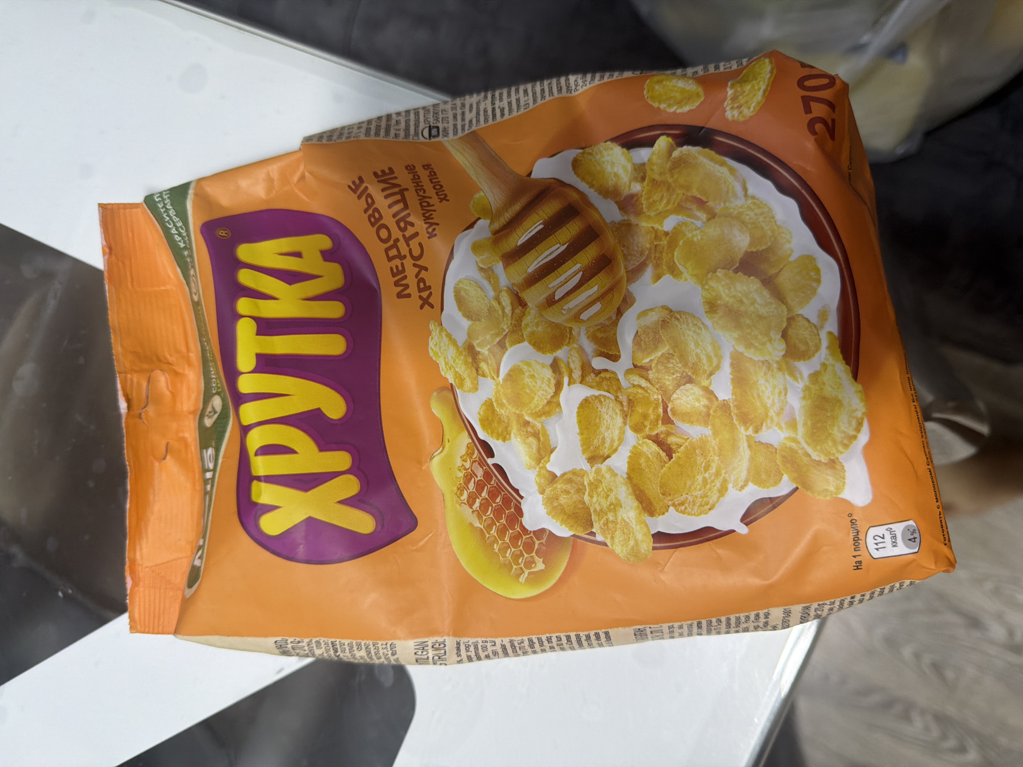The image shows a close-up of an orange cereal box, likely packaged corn flakes. The brand name "ХРУТКА" is prominently displayed in large yellow lettering within a purple outline. Below this, text in Russian reads "МЕДОВЫЕ ХРУСТЯЩИЕ хлопья," which translates to "HONEY CRISPY flakes." The packaging features an illustration of a bowl of cereal with milk, honeycombs, and a honey dipper. On the bottom right corner of the box, the number "270" is visible, possibly indicating the weight in grams. Nutritional information is also partially visible, showing "112 ккал" (112 kcal) per serving and "4%". The cereal box is placed on a surface that appears to be a light-colored countertop or table, with some shadows and reflections visible. There are no people or specific city landmarks visible in the image.