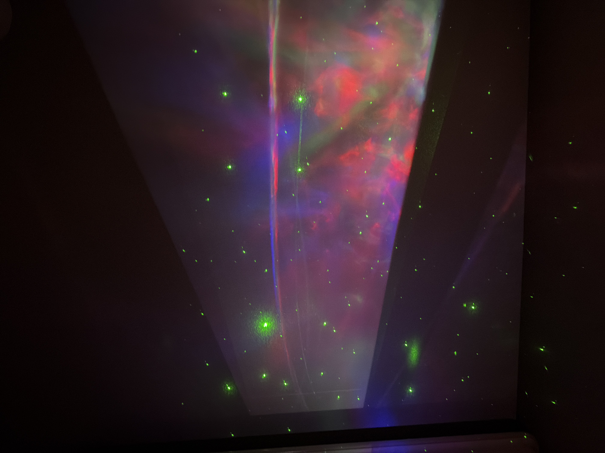 The image shows a dark room interior, likely a bedroom, with projections cast onto the ceiling and walls. The projections include diffuse patterns of red, blue, and green light, resembling a nebula or galaxy effect. In addition, numerous small, focused green laser dots are scattered across the surfaces. The light sources are not visible in the image, but they are presumably located outside the frame. The lighting conditions suggest nighttime. The presence of projections and laser lights indicates the use of a lighting device for recreational or aesthetic purposes. The location is not discernible based on the provided image and therefore no information about it can be provided.