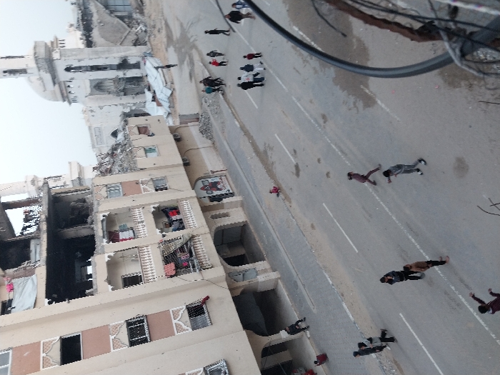 A scene of destruction, likely in a war zone. A damaged building exposes interiors. A mosque with a partially collapsed dome stands nearby. People are scattered along a wide, empty street, some lying prone, suggesting casualties or seeking cover. Cables are visible in the upper right. The overall impression is one of conflict and its aftermath.