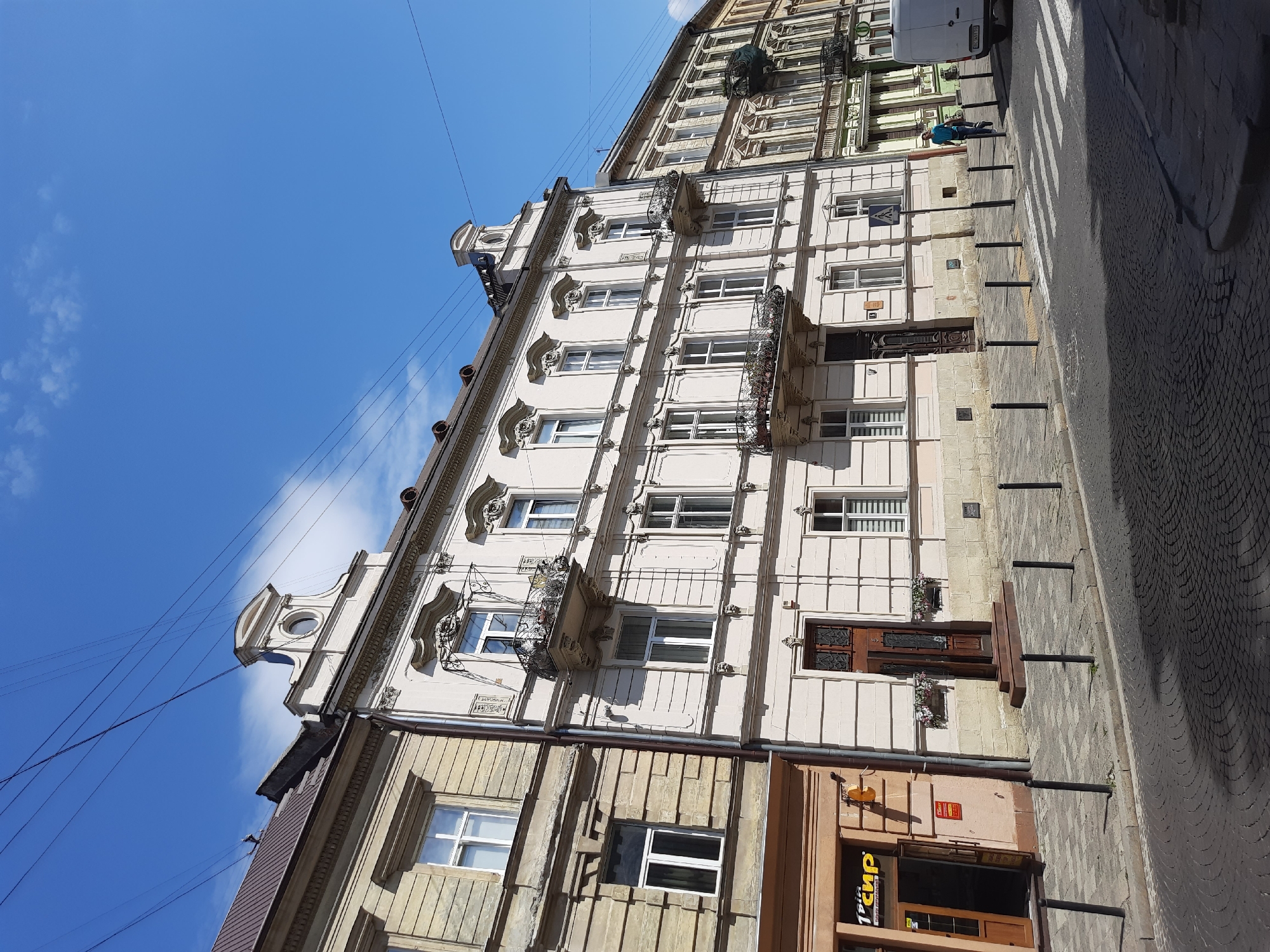 This image depicts a street scene in Lviv, Ukraine, featuring a row of European-style buildings with ornate facades and arched windows. The architecture suggests a historical district. The street is paved with cobblestones, and a pedestrian crossing is visible. Overhead, a network of wires crisscrosses the blue sky, dotted with wispy clouds. A person in a blue jacket is walking across the street near a white van. The buildings display a variety of architectural details, including balconies with intricate ironwork, decorative molding, and mansard roofs. One building on the left has a sign that reads "1 пир". The overall impression is of a sunlit day in an old European city.