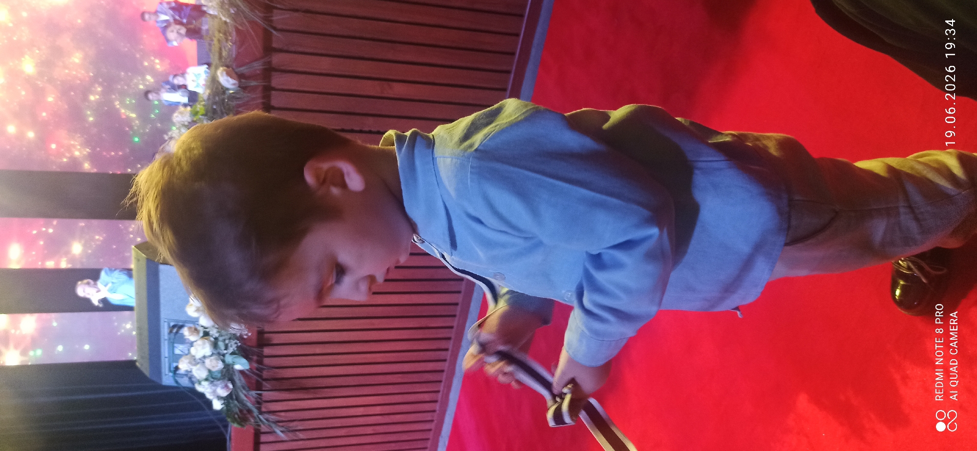 A young boy, seen from the chest up and side profile, stands on a red carpet in front of a wooden paneled wall. He is wearing a light blue collared shirt and light-colored pants. The boy is holding a ribbon with black and white stripes in his hands, appearing to be tying or examining it. In the background, on a stage, several people are visible, along with a large speaker and a floral arrangement. The stage is illuminated with a colorful, sparkling light display. The scene appears to be an indoor event, possibly a performance or ceremony.