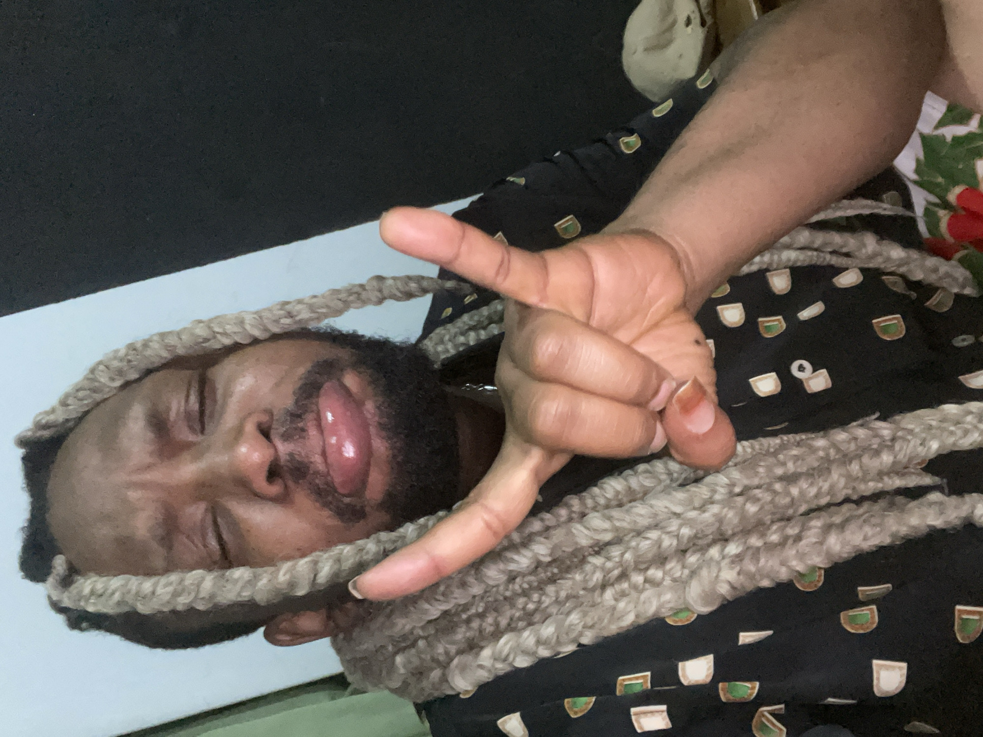 The image shows a person with braided hair lying down, possibly in an indoor setting. The person's hand is raised, making a hand gesture. The person is wearing a patterned shirt. Without additional information, I cannot confirm the location in the city of Jos, Nigeria.