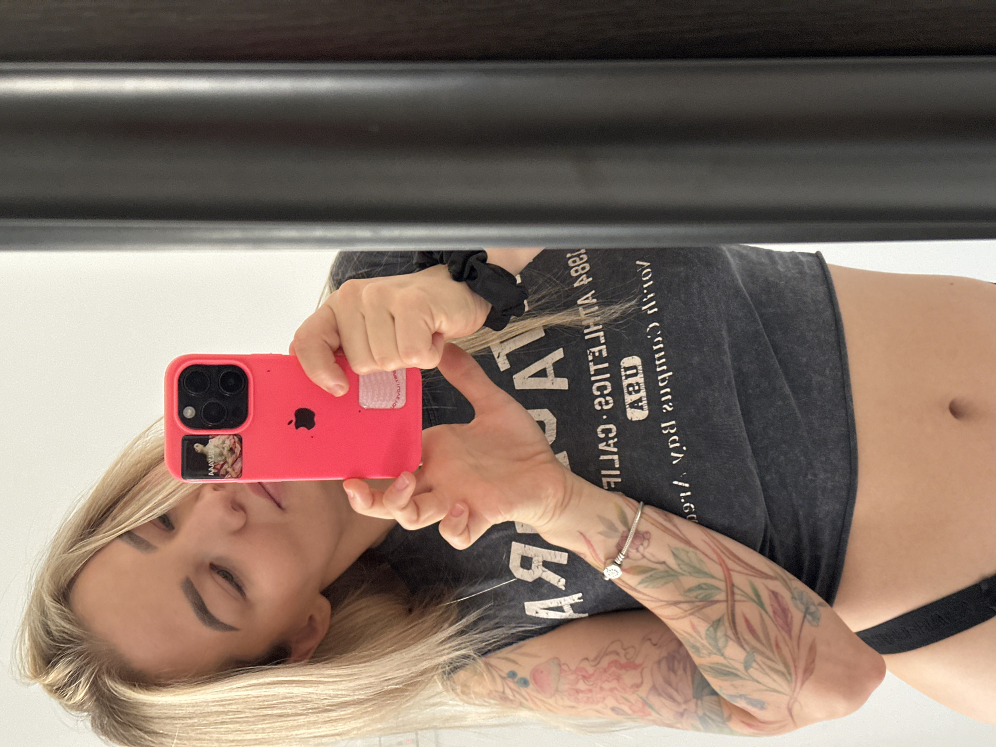 The media file presents a mirror selfie of a young woman, currently oriented on its side, with her head positioned on the left of the frame. She has blonde hair and a relaxed expression, with her eyes partially closed.
She holds a vibrant pink iPhone case, which features a cutout revealing the phone's metallic Apple logo. Below this, a small sticker with an artistic motif, possibly a classical painting or floral design, is visible on the case. A black scrunchie is worn on her right wrist, while her left wrist is adorned with a silver bracelet carrying a small, intricate charm. Both of her arms are extensively decorated with colorful floral and foliage tattoos.
She is wearing a dark grey cropped t-shirt, upon which reversed text is visible due to the mirror's reflection. The text clearly reads "CALIFORNIA VINTAGE 1984". A black bra strap is visible below the hem of her shirt, revealing her midriff and navel. The background is a reflection of a bright, light-colored surface, possibly a wall or ceiling, topped by a dark, horizontal bar. This scene is set in Budva, Montenegro.