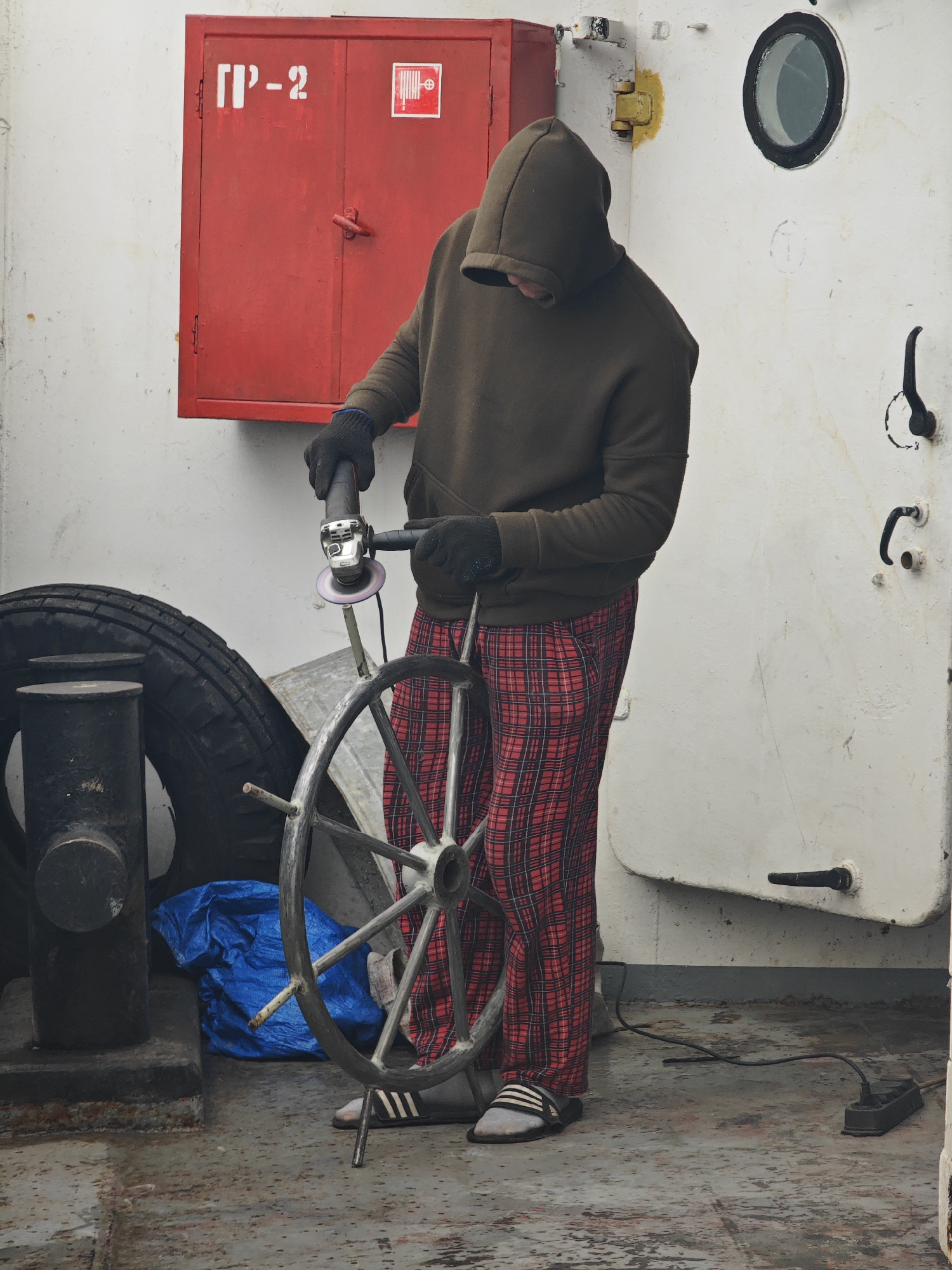 A person wearing a dark green hooded sweatshirt, black gloves, and red and black plaid pants is working on a metal ship's wheel with a handheld grinder. The person's face is obscured by the hood. The grinder is being held with both hands and is actively grinding the metal wheel. To the left of the person, there is a large black tire and a black bollard. A bright blue tarp is visible behind the tire. In the background, there is a red fire extinguisher cabinet mounted on a white wall. To the right of the person, there is a white circular porthole with a black frame and a black handle on the wall. The scene appears to be on the deck of a ship or a similar industrial setting. The ground is a weathered metallic surface with some stains. The image is taken from a medium distance, capturing the upper body of the person and the immediate surroundings. The overall lighting is neutral. This scene is not located in the city of Ермолаево, Russia.