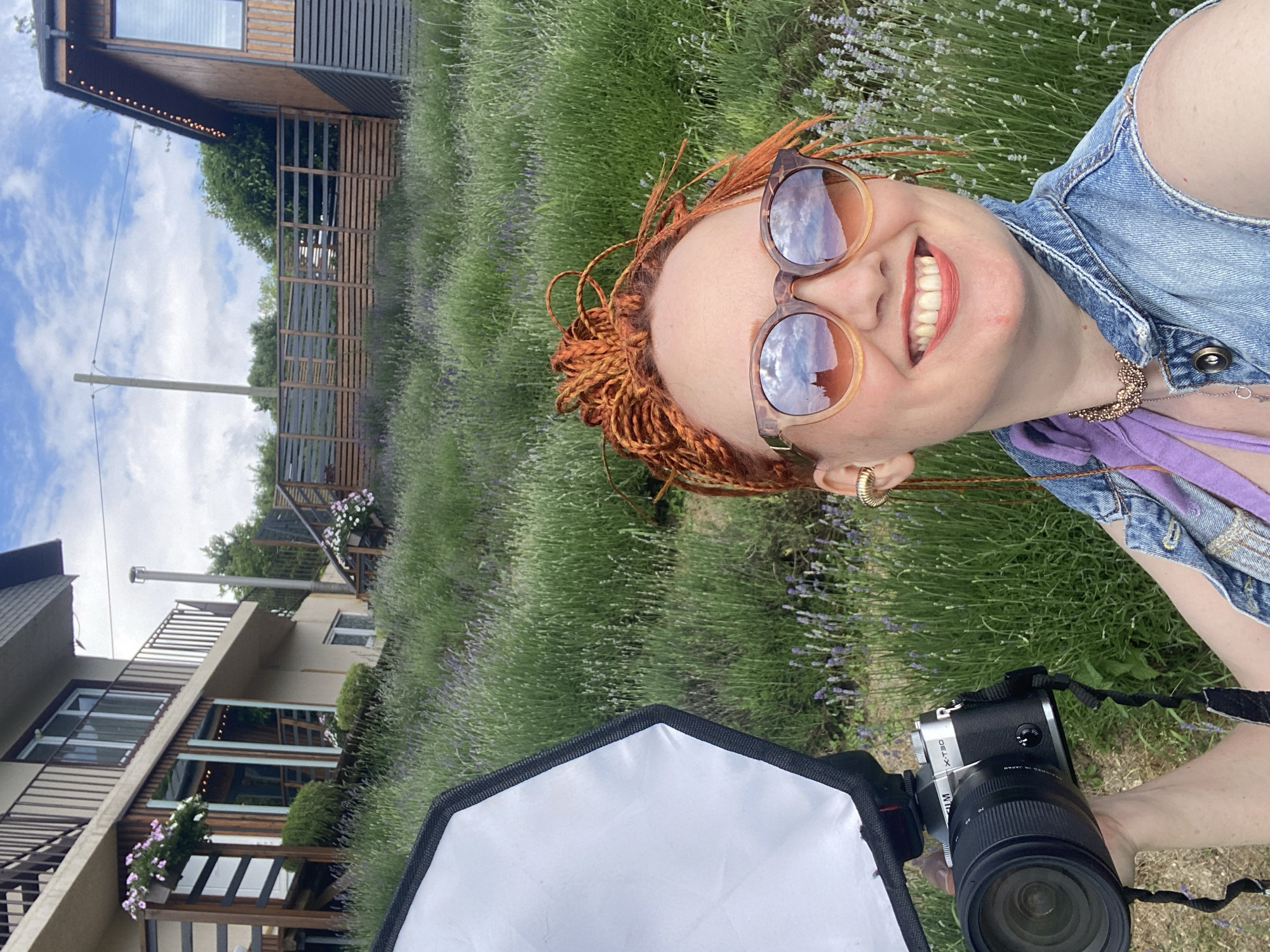 An outdoor, medium shot captures a woman taking a selfie in a field of lavender. The woman has bright orange braided hair and is wearing sunglasses, a denim vest over a purple top, and a gold necklace. She is smiling and holding a camera with a softbox attachment. In the background, there are two buildings with wooden accents and a fence, set against a backdrop of lush green vegetation and a cloudy sky. The location is Verkhneye Uchdere, Russia, suggested by the overall atmosphere and vegetation.