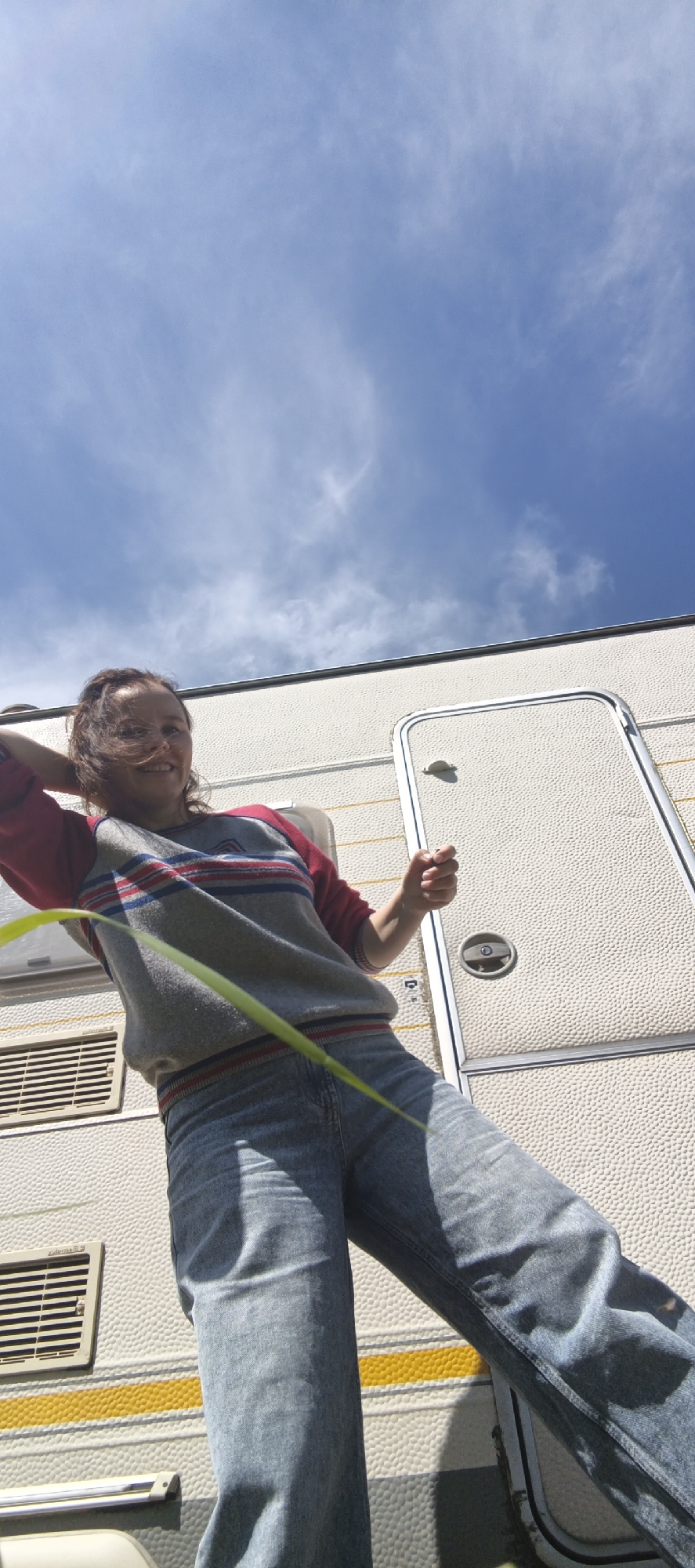 An outdoor, low-angle, vertical shot captures a young woman from the waist down, standing in front of a white recreational vehicle (RV) on a bright, cloudy day. The RV, which occupies the right and lower portion of the frame, has a textured surface and a closed door on the right. A yellow stripe runs horizontally across the RV's lower exterior. In the upper portion of the image, a blue sky with scattered white clouds is visible. The woman is wearing a gray sweatshirt with red and blue stripes across the chest and blue jeans. A thin, green blade of grass extends diagonally across the foreground, obscuring part of the woman's torso and legs. The image appears to have been taken in a natural outdoor setting, possibly a campsite, but no specific location within Chaikovsky, Russia, is identifiable from the visual information provided.