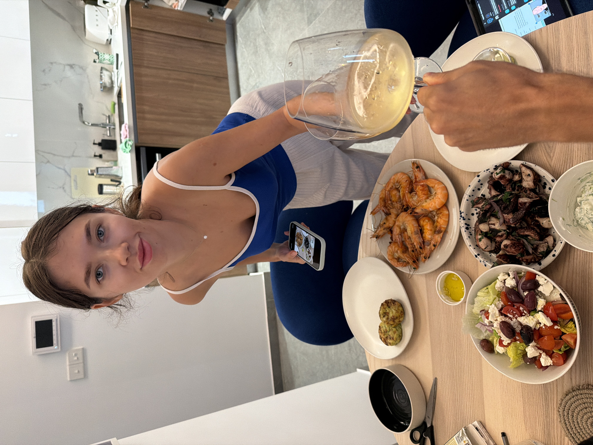 A young woman in a blue and white tank top stands next to a table, holding a smartphone in her left hand, seemingly photographing or recording a video of the food spread on the table. She is tilting a pitcher of a light-colored beverage, presumably wine, into a glass held by another person whose hand is visible in the top right of the frame. The table contains a variety of dishes: a plate of grilled shrimp, a plate of grilled octopus, a Greek salad with olives and feta cheese, a small dish of olive oil, and a plate with what appears to be two small patties. A black bowl, scissors, and other items are also on the table. The scene is indoors, likely in a dining room or kitchen, given the presence of cabinets and countertops in the background. The location is not visibly identifiable as Livadia Larnakas, Cyprus based solely on the image content.