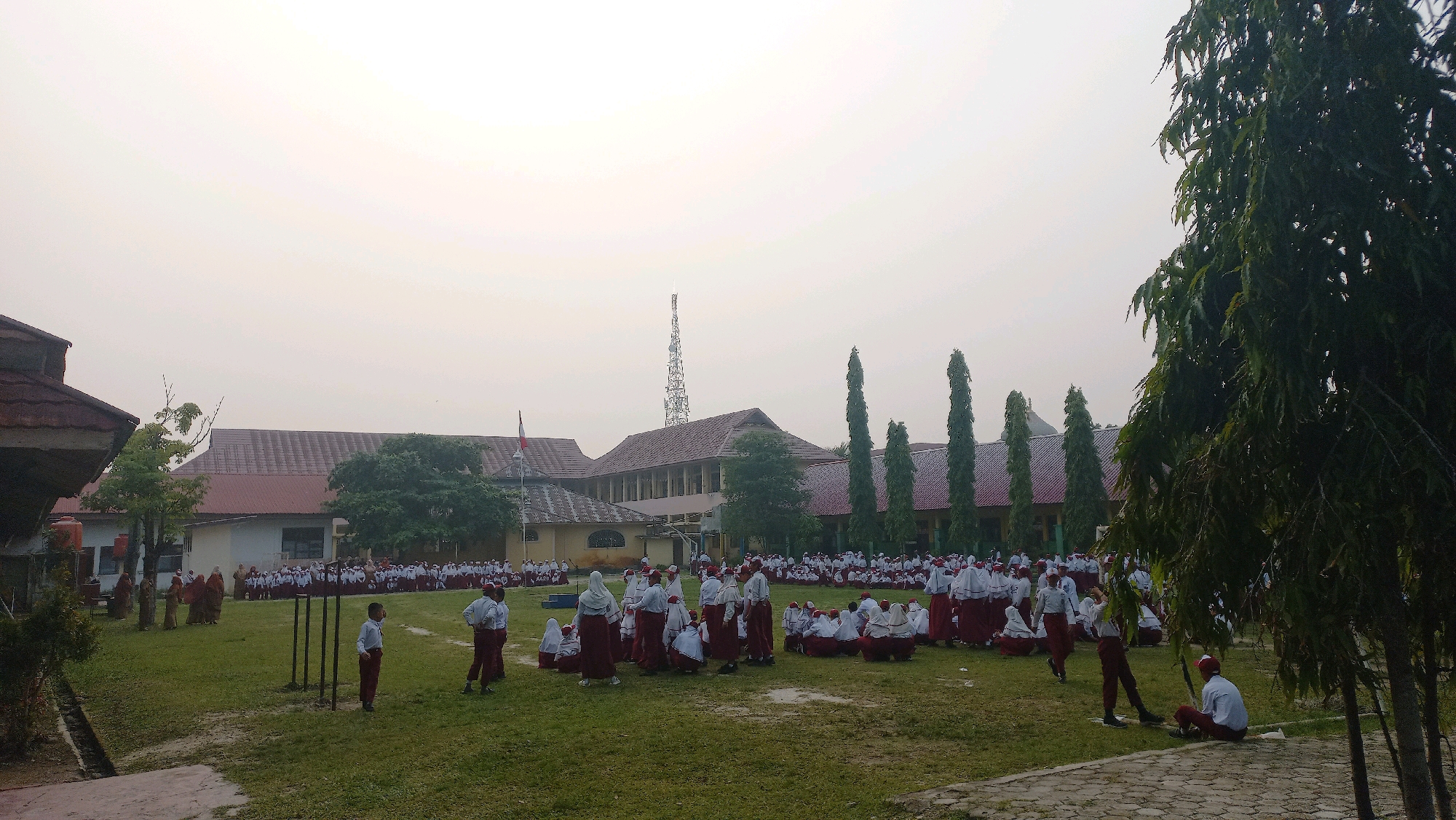 The image depicts an outdoor scene at a school in Bangkinang, Indonesia, likely during a morning assembly or activity. A large group of children, dressed in school uniforms of white shirts and maroon skirts or pants, are gathered on a grassy field. Some children are standing in rows, while others are sitting on the ground, suggesting an organized activity. In the background, a long, multi-story school building with red tiled roofs is visible. Trees, including a prominent large tree on the right side of the frame, line the field and surround the building. A telecommunications tower can be seen in the distance above the school building. The sky is overcast, contributing to a soft, diffused light across the scene. A few adults, also in traditional attire, are present among the children, possibly supervising. The overall atmosphere appears to be one of a typical school day in a tropical or subtropical environment.