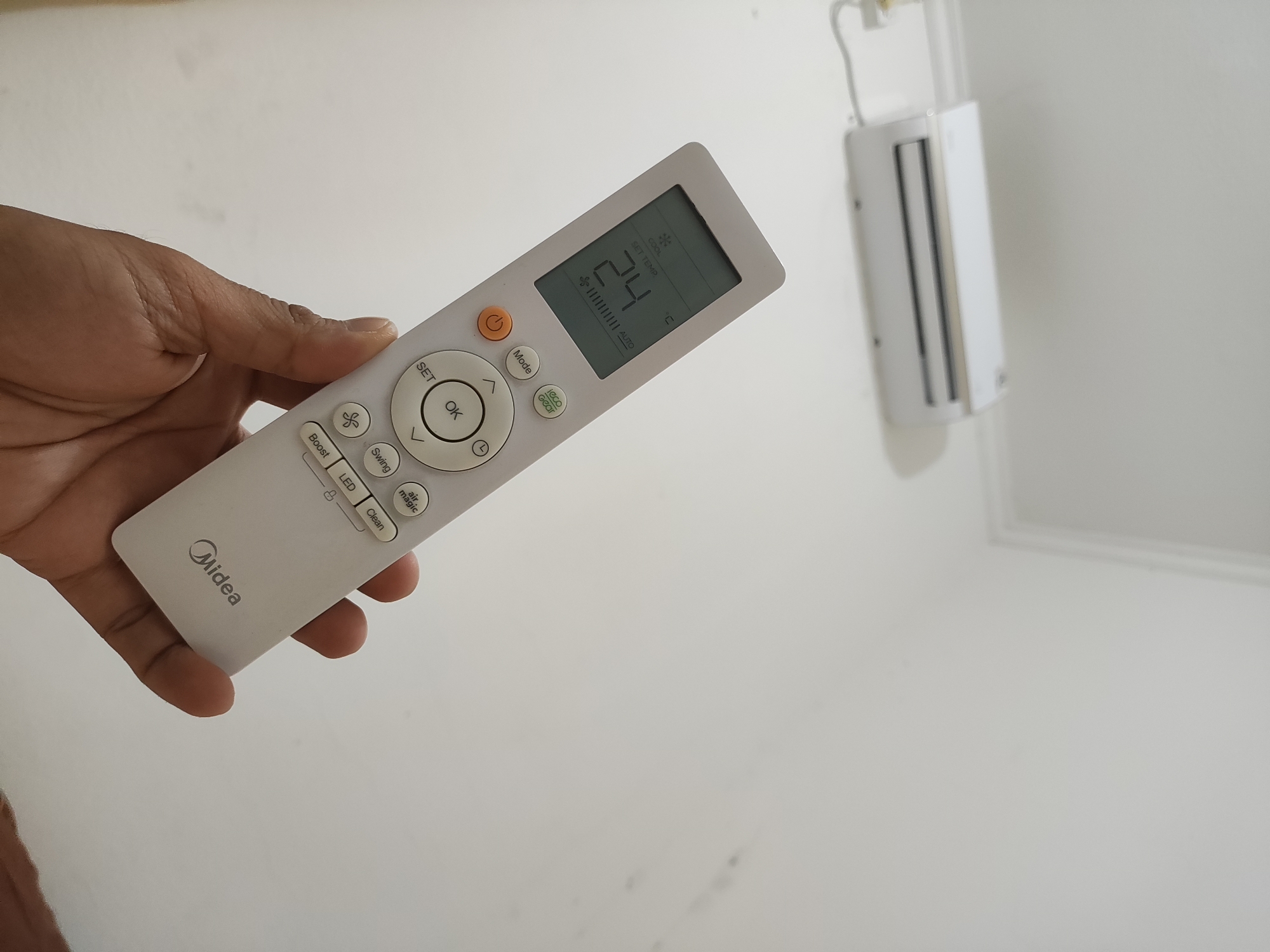 In a well-lit indoor environment, a person's hand is holding a white remote control, likely for an air conditioning unit. The remote has a digital display showing "24" degrees Celsius, indicating a set temperature. Various buttons are visible on the remote, including "SET", "OK", "Mode", "Boost", "Swing", "LED", and "Clean". To the right and slightly out of focus, a white air conditioning unit is mounted on a wall. The overall scene suggests a typical domestic or office setting. The image does not contain any elements that would allow for the identification of a specific location in Wanajaya, Indonesia.