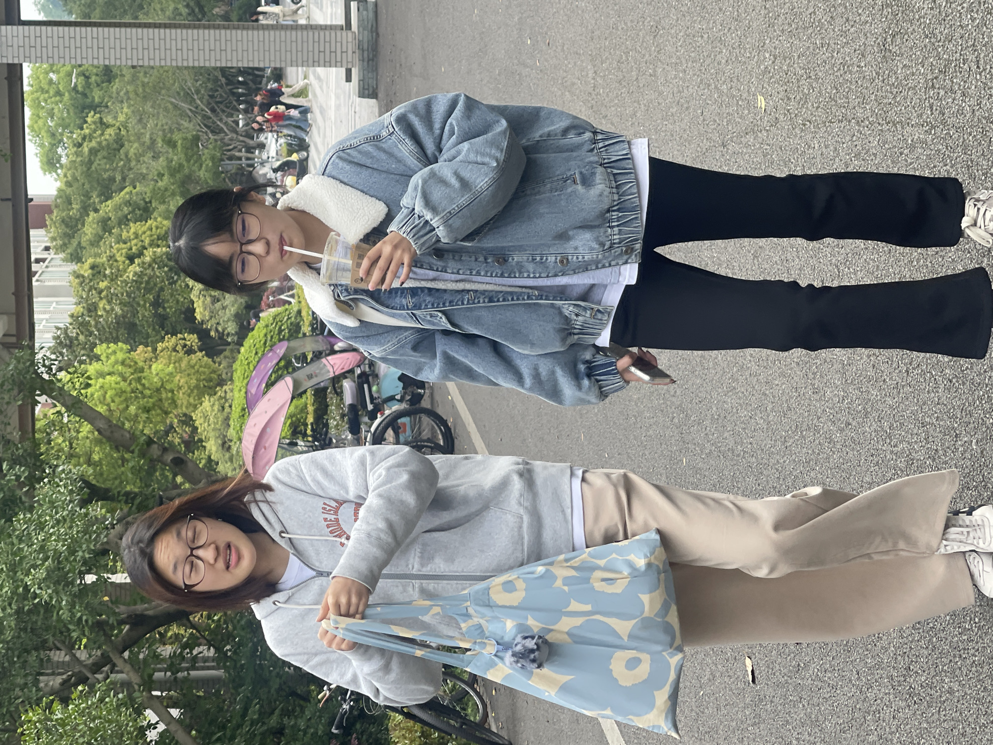 Two women stand on a paved area, likely a sidewalk, surrounded by lush green trees and parked bicycles. The woman on the right wears a denim jacket with a white shearling collar, black pants, and sneakers. She is holding a cup with a straw and a mobile phone. The woman on the left wears a gray hoodie, beige pants, and sneakers, holding a blue floral pattern tote bag with a pom-pom detail. The scene suggests a college or university environment in the Chongqing High-tech Zone in China.
