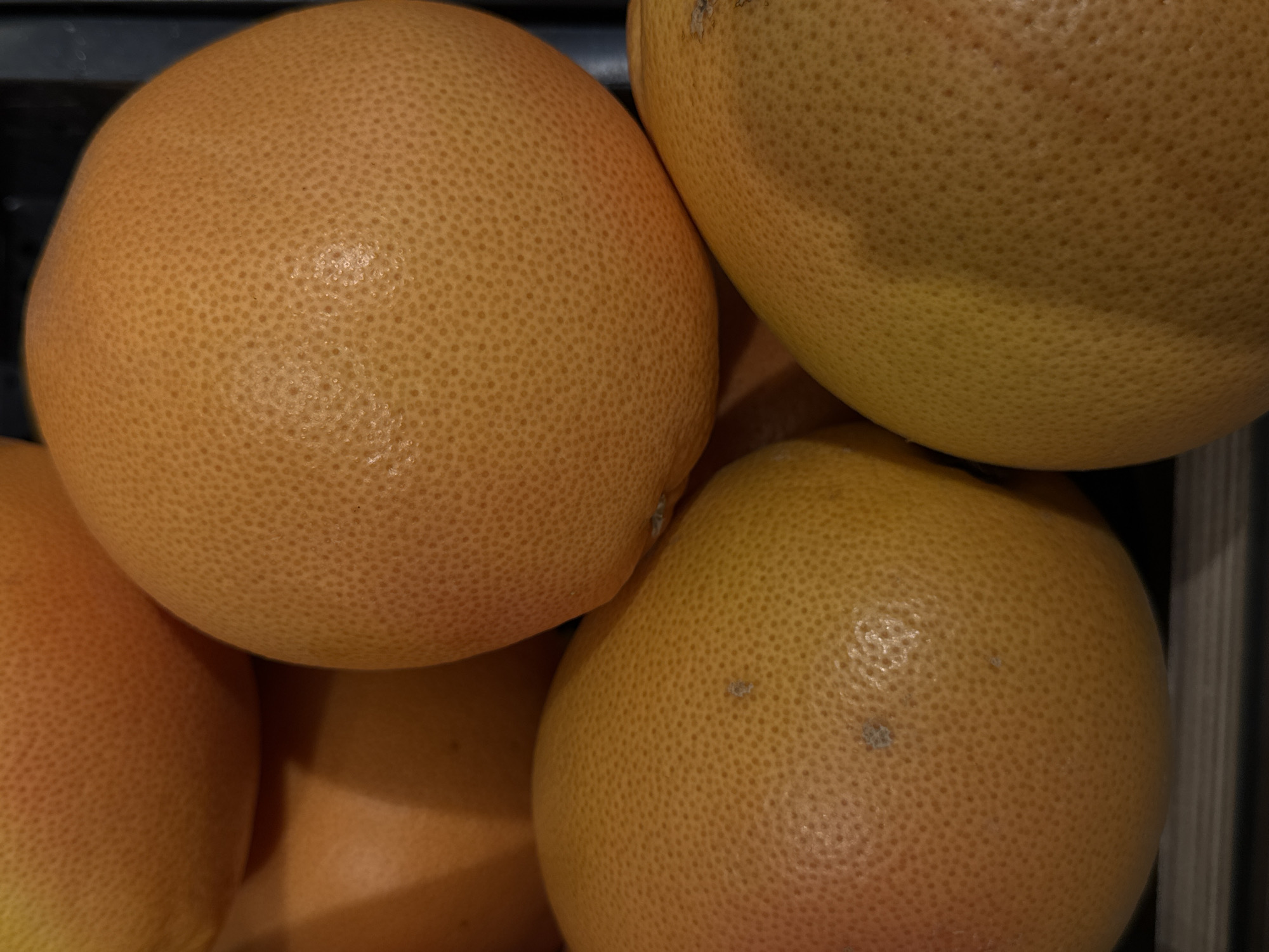 The media file is a close-up, high-angle shot of four grapefruits. The grapefruits are the primary subjects, filling the entire frame. They are round and have a textured peel with visible pores. The color of the grapefruits ranges from a pale yellow to a light orange-pink hue. Some slight imperfections, like small light-colored spots, are visible on the peel of a few of the fruits. The lighting is somewhat dim, casting soft shadows and highlighting the texture of the fruit. The background is dark and out of focus, with hints of what appear to be dark shelving or a container. There are no people or any discernible activity taking place. The image appears to be taken indoors, likely in a grocery store or home setting. No text is visible in the image. Given the location context of Moscow, Russia, these could be grapefruits purchased or displayed in a Russian market or home.