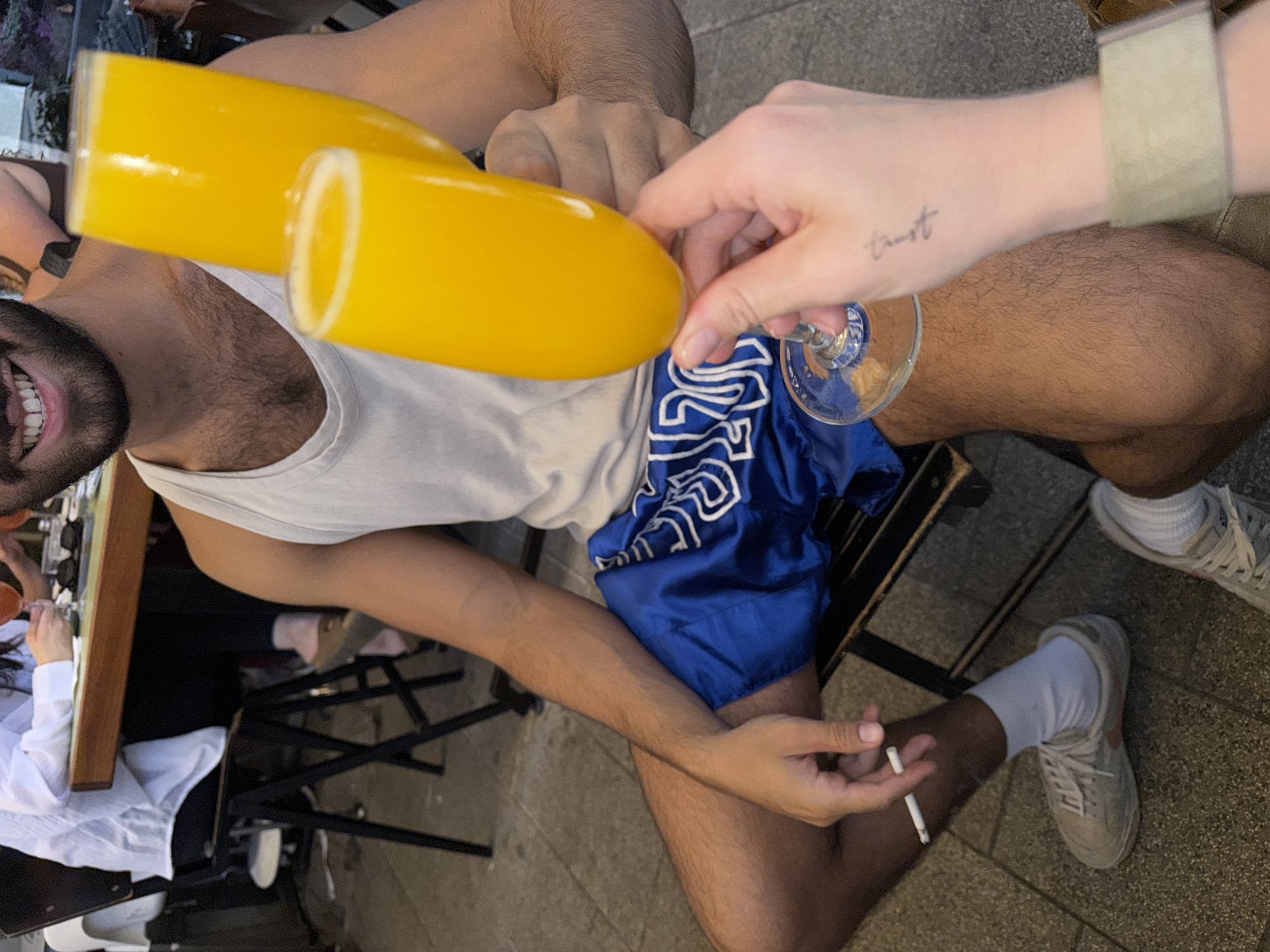 A photograph taken from an overhead perspective captures two individuals seated at an outdoor cafe, potentially in Tel Aviv, Israel. One person, visible from the chest up, is wearing a white tank top. This individual, a man with dark hair and a beard, holds a glass filled with a vibrant yellow liquid, presumably orange juice. Another hand, adorned with a wristwatch and a tattoo reading "trust," holds a similar glass. The man is seated on a black chair, wearing blue shorts. He holds a cigarette in his hand. Another person is visible to the left, partially obstructed by a table. The pavement suggests an urban setting.