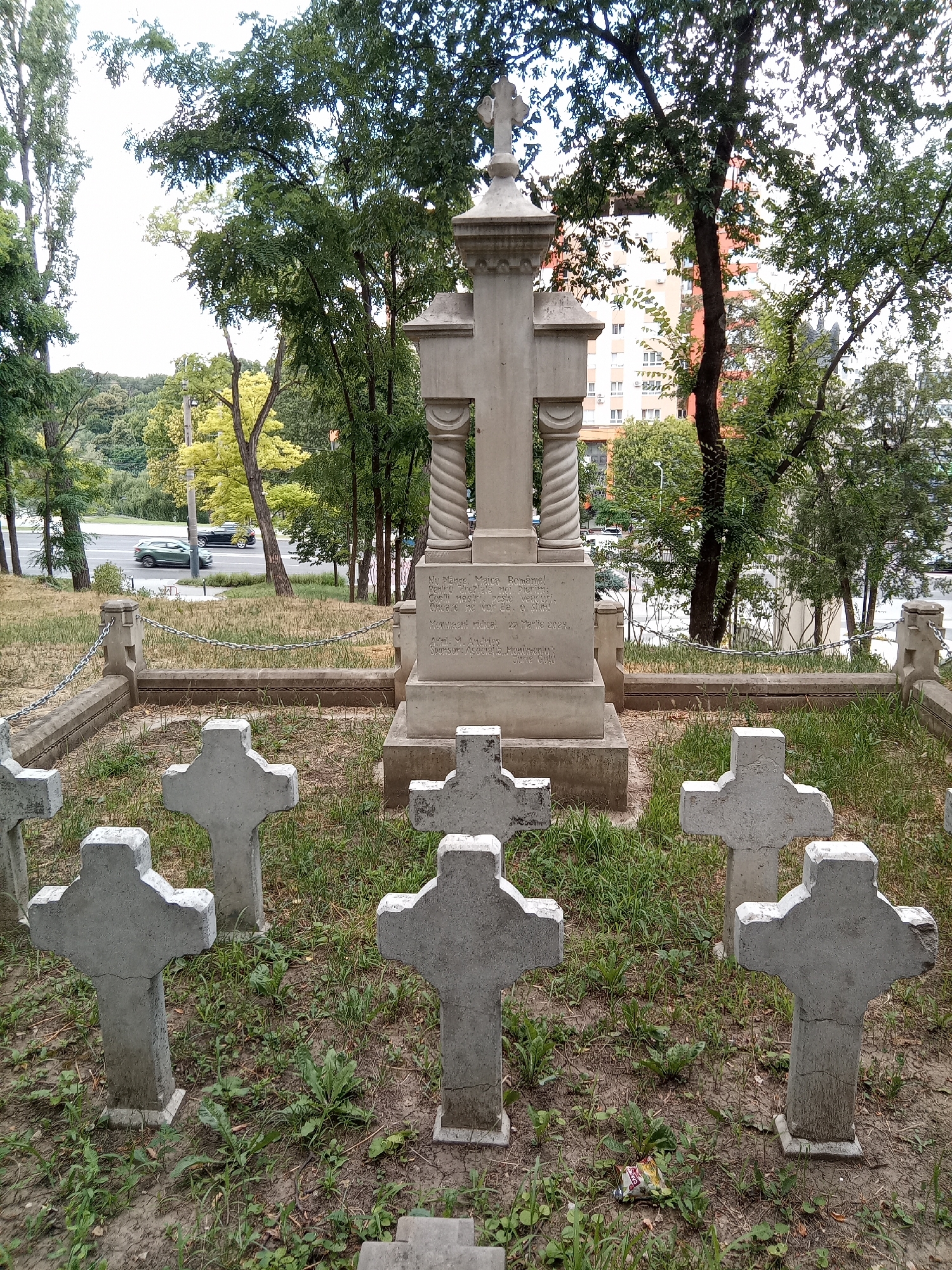 The image shows a monument and several simple cross-shaped grave markers in a cemetery. The monument is a stone structure with a cross at its peak, flanked by two twisted columns. The grave markers are made of concrete and are arranged in a semi-circular pattern around the monument. The cemetery is overgrown with grass and weeds, and a chain-link fence surrounds the monument. In the background, there are trees, buildings, and a street with cars. The scene appears to be in a public park or a cemetery located in a city.
The location is the "Monument to the Heroes of the Uprising of the Grivitsa" in Chișinău, Moldova. This monument commemorates the Romanian soldiers who died in the battle of Grivița during the Russo-Turkish War of 1877-1878.