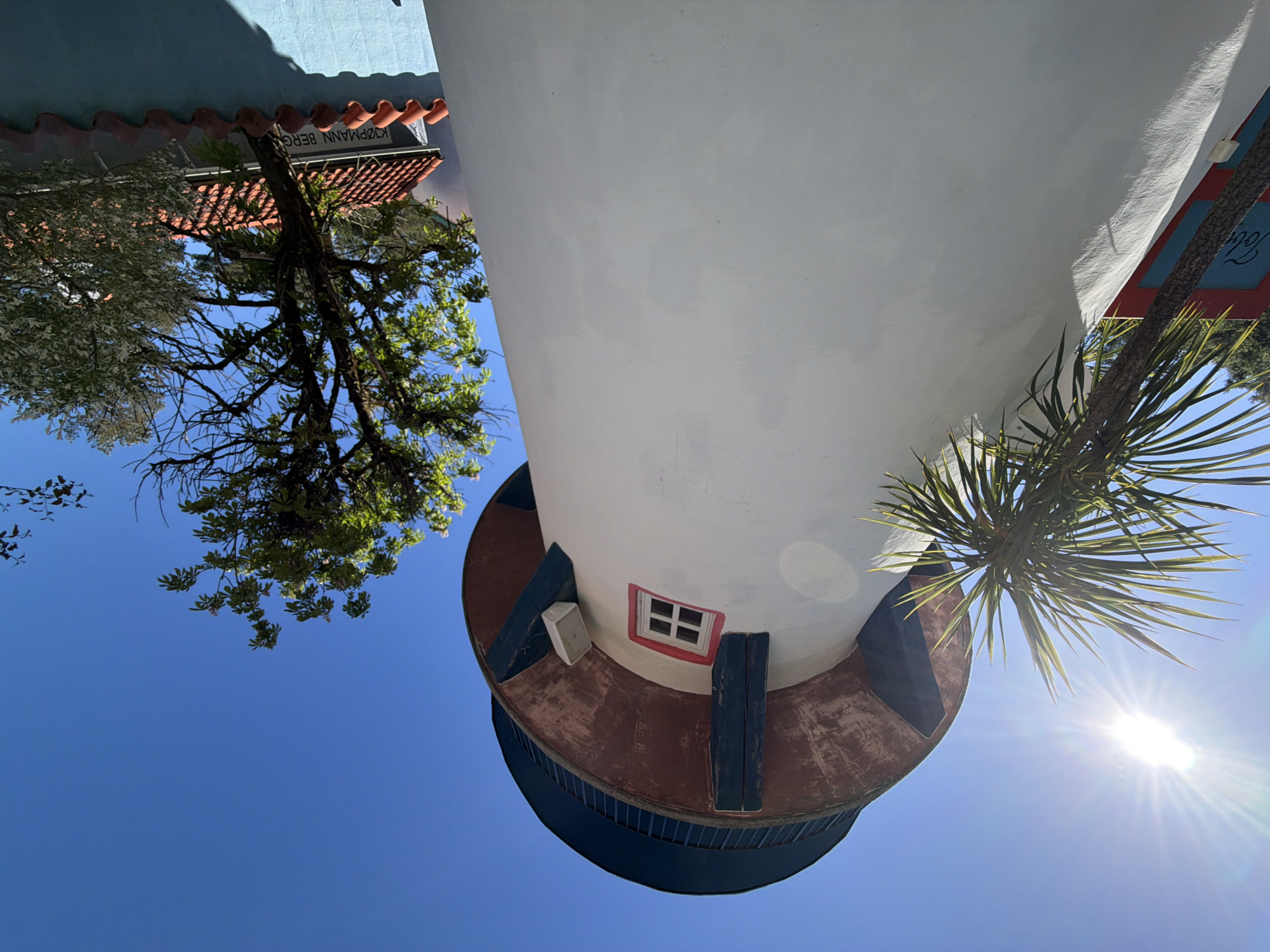 This image depicts an outdoor scene under a clear blue sky with intense sunlight, evidenced by a prominent sunburst lens flare in the upper left. The central and dominant feature is a tall, white cylindrical building, resembling a water tower or lighthouse, viewed from a low-angle perspective. The building's upper portion consists of a wide reddish-brown band supporting a dark blue top section, which features a railing. Several dark blue, vertical support beams are visible beneath the reddish-brown band. On the white facade, a small, square window with a white grid frame is accented by a red border. To the right of this window, a small, off-white rectangular object is mounted on the wall.
On the left side of the frame, a tall, palm-like plant with long, pointed green leaves partially obscures the building. On the right, a tree with vibrant green leaves and white blossoms is visible against the sky, with another tree featuring lighter foliage further right. In the lower right quadrant, a building with a reddish-orange tiled roof is partially visible. Below its roofline, a light grey wall displays white lettering that reads "KJØPMANN BERG". Beneath this, a light blue-green wall with a wavy, reddish-brown tiled capping is present, casting a shadow. A partial red and blue sign with white script text, reading "Tol", is faintly visible in the lower left, behind the palm-like plant.
Based on the identifiable text "KJØPMANN BERG", the location is in Kristiansand, Norway, specifically in the vicinity of Kjøpmann Berg Båt og Motor, which is located in the city's harbor area. The additional partial text "Tol" suggests proximity to a customs house or harbor facility. No human subjects are present in the image.