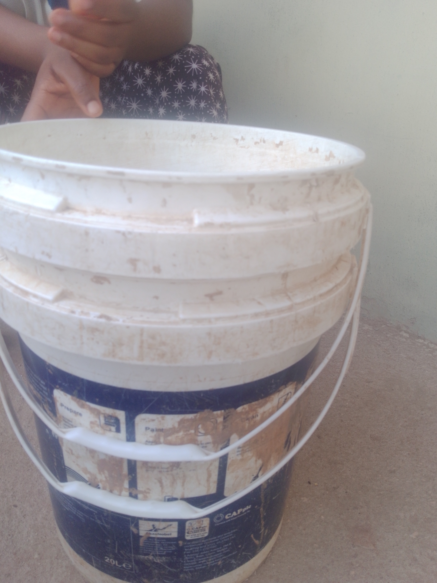 A person's hands are visible at the top of the frame, appearing to be interacting with a dirty, white plastic bucket. The bucket has a blue and white label that is partially obscured by dirt. The bucket is sitting on a light brown, textured surface, possibly concrete. The background is a light-colored, textured wall. The overall scene appears to be outdoors, possibly in an urban or semi-urban environment, but no specific location details can be identified.
