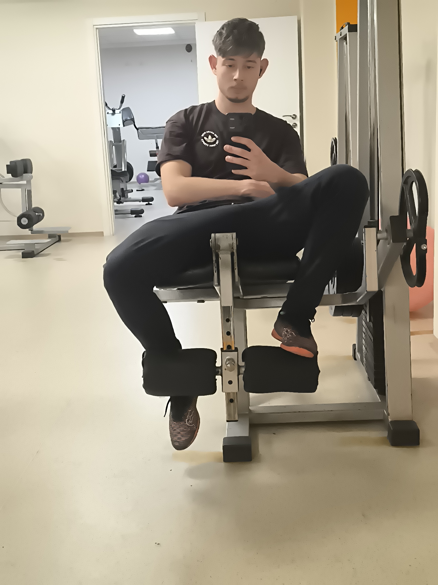 A young man is sitting on a leg extension machine in a gym setting. He is wearing a black Adidas t-shirt and black sweatpants. He is holding a smartphone in his hand, seemingly looking at the screen. The gym has standard equipment and a light-colored floor. The location is potentially in Sofia, Bulgaria, but no landmarks confirm this.