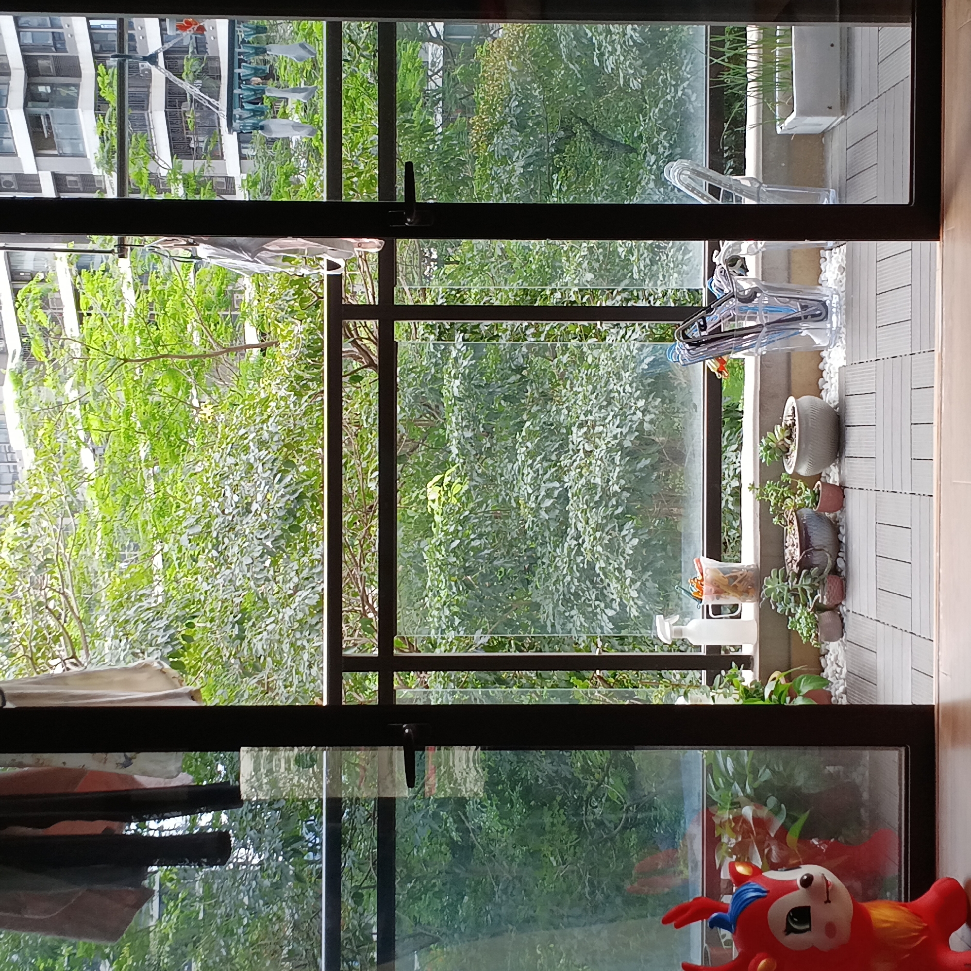 A view from inside a building in Shunde District, China, looking through an open balcony door. Outside the balcony door, lush green trees and foliage are visible. On the balcony are potted plants and gardening supplies, including a spray bottle, containers, and plant pots. A red toy with a cartoon face sits in the foreground, inside the building. In the distance, other buildings can be seen. Laundry is hanging on a clothesline outside a building in the distance.