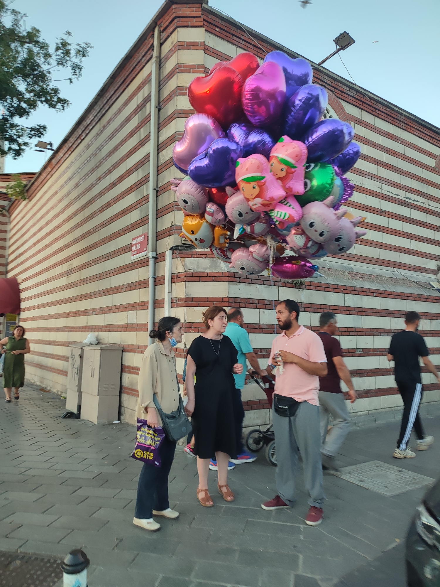 In Üsküdar, Turkey, a diverse group of people are gathered on a pedestrian sidewalk. Prominently displayed is a large bunch of colorful balloons, including heart-shaped ones and cartoon characters, tethered to a vendor's cart. A man in a pink shirt is holding a drink and speaking with a woman in a black dress, while another woman in a beige jacket looks on. Behind them, several other individuals are walking or standing, some obscured by the balloons. The background features a building with a distinctive red and white striped facade, typical of many structures in the historic district of Üsküdar. The scene appears to be set in the late afternoon or early evening, with soft natural light illuminating the area.