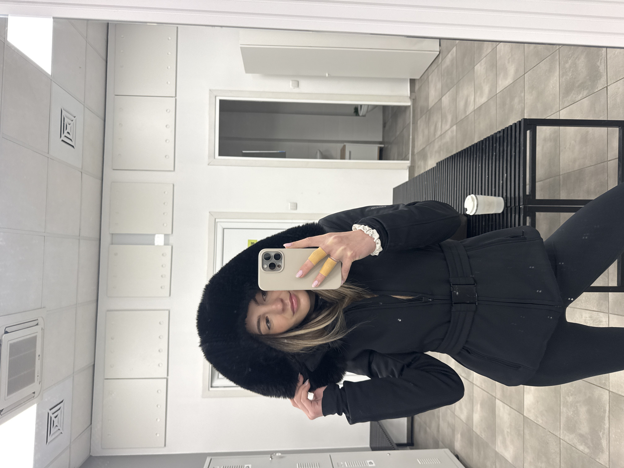 The image shows a woman taking a selfie in a room that appears to be a changing or locker room. The room has white tile walls, metal lockers, and a bench with a coffee cup on it. A door can be seen in the background, as well as a glimpse of a room beyond. The woman is wearing a black, hooded winter jacket and black leggings. She is holding a cell phone in her hand, obscuring some of her face. It's not possible to determine if the location is Sofia, Bulgaria, without further information.