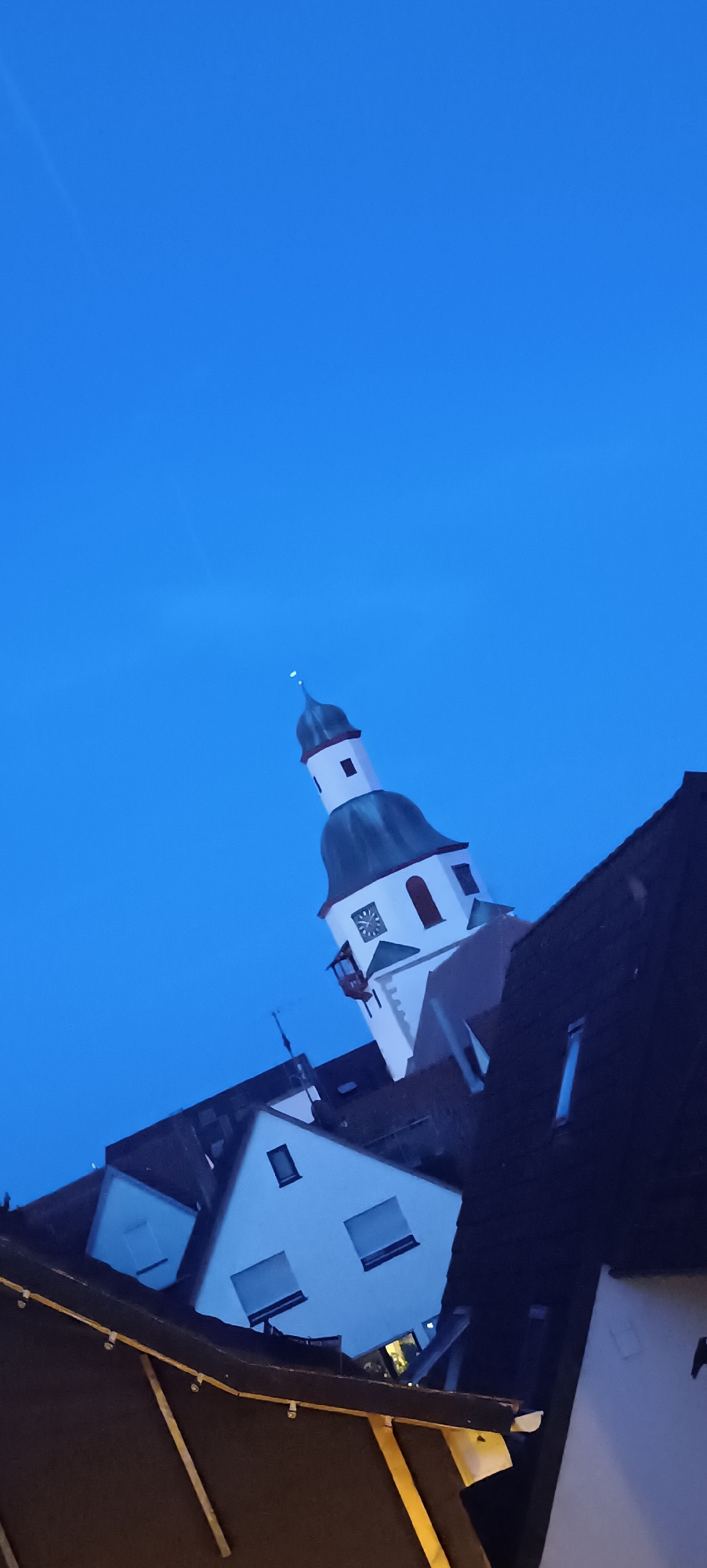 This is a vertical, low-angle, nighttime photograph of the tower of the Protestant Stadtkirche Winnenden in Winnenden, Germany. The sky is a deep, clear blue, indicative of twilight or night. The church tower is white with a dark green, conical roof topped with a small spire. A clock face is visible on the tower, along with a small balcony and some windows. In the foreground and to the left, several rooftops of houses are visible, with dark brown or black tiles. Some of these houses are modern, with white facades and rectangular windows. The overall impression is of a European town seen from below at dusk or dawn.