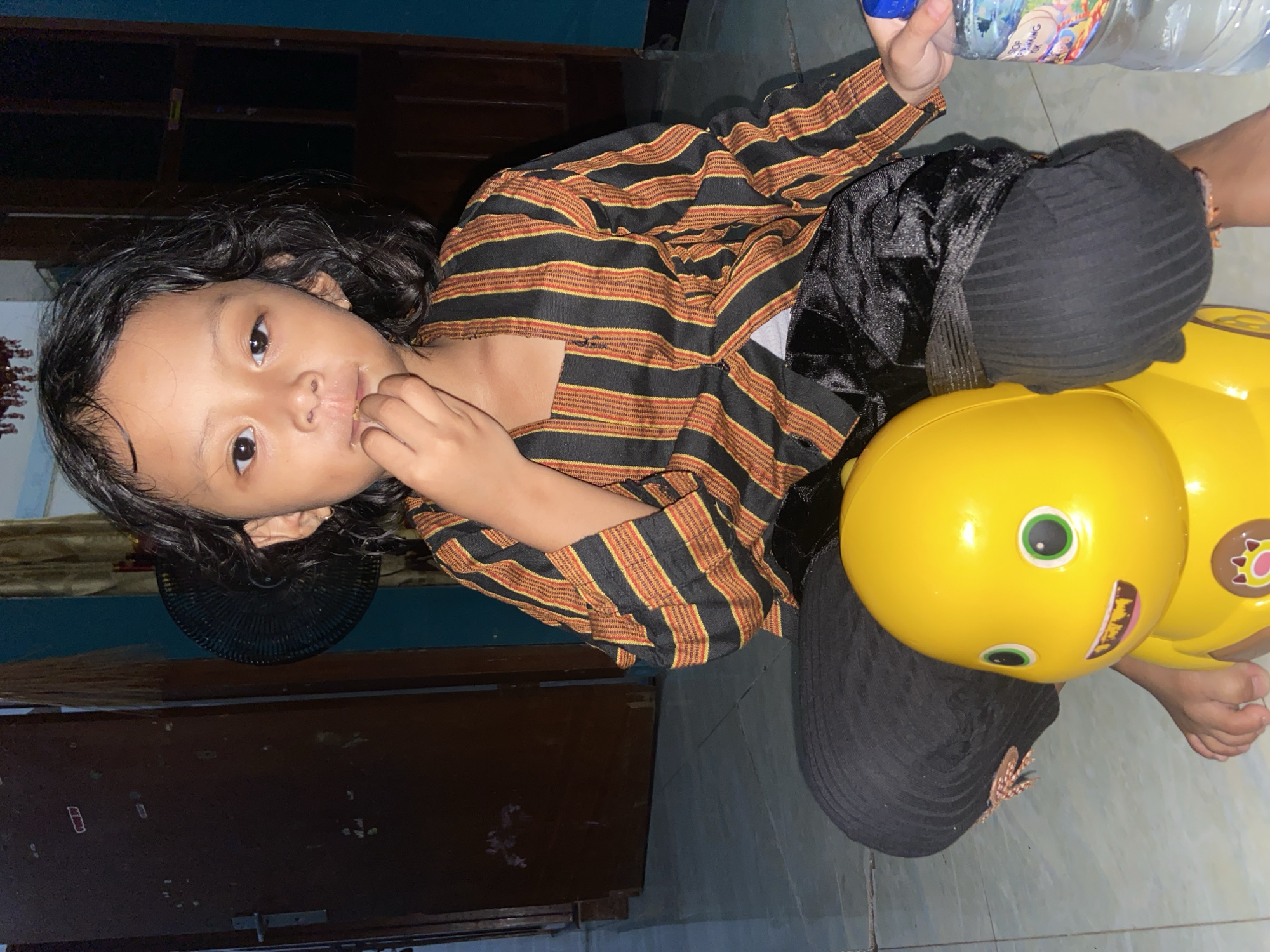 A child in Gelam, Indonesia, sits on a yellow toy, possibly a ride-on toy. The child wears a striped brown and black shirt, and a dark garment is draped over their lap. The child is holding a clear plastic water bottle. The environment includes a dark-colored wooden cabinet, a fan, and tiled flooring.