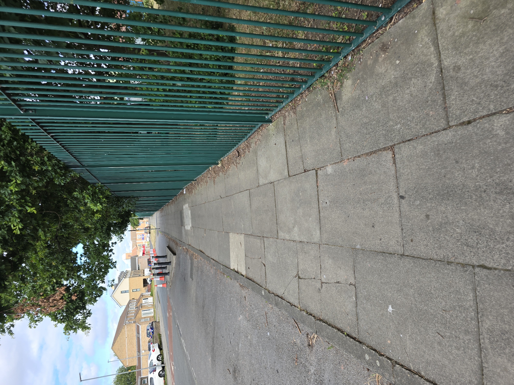 The image shows a sidewalk next to a road and a green metal fence. The sidewalk is made of concrete slabs. The road has white lines and traffic cones on it. In the background, there are houses and trees. The sky is blue with some clouds. The scene appears to be in a residential area of Stockton-on-Tees, United Kingdom.
