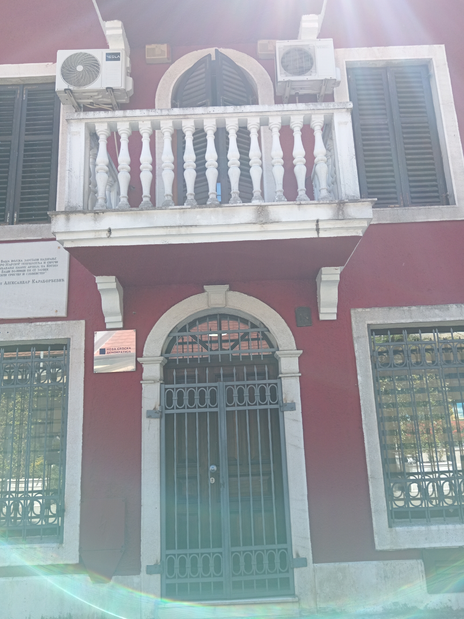 The image depicts the facade of a maroon-colored building in Podgorica, Montenegro. The building features a balcony with white balusters, two air conditioning units, windows with metal grilles, and a metal gate over the entrance. A plaque with Serbian text is mounted on the wall to the left of the entrance. A sign reading "NOVA SRPSKA DEMOKRATIJA" (New Serbian Democracy) is placed between the plaque and the gate. The building appears to be located in a urban environment. There are no people directly visible in the frame.