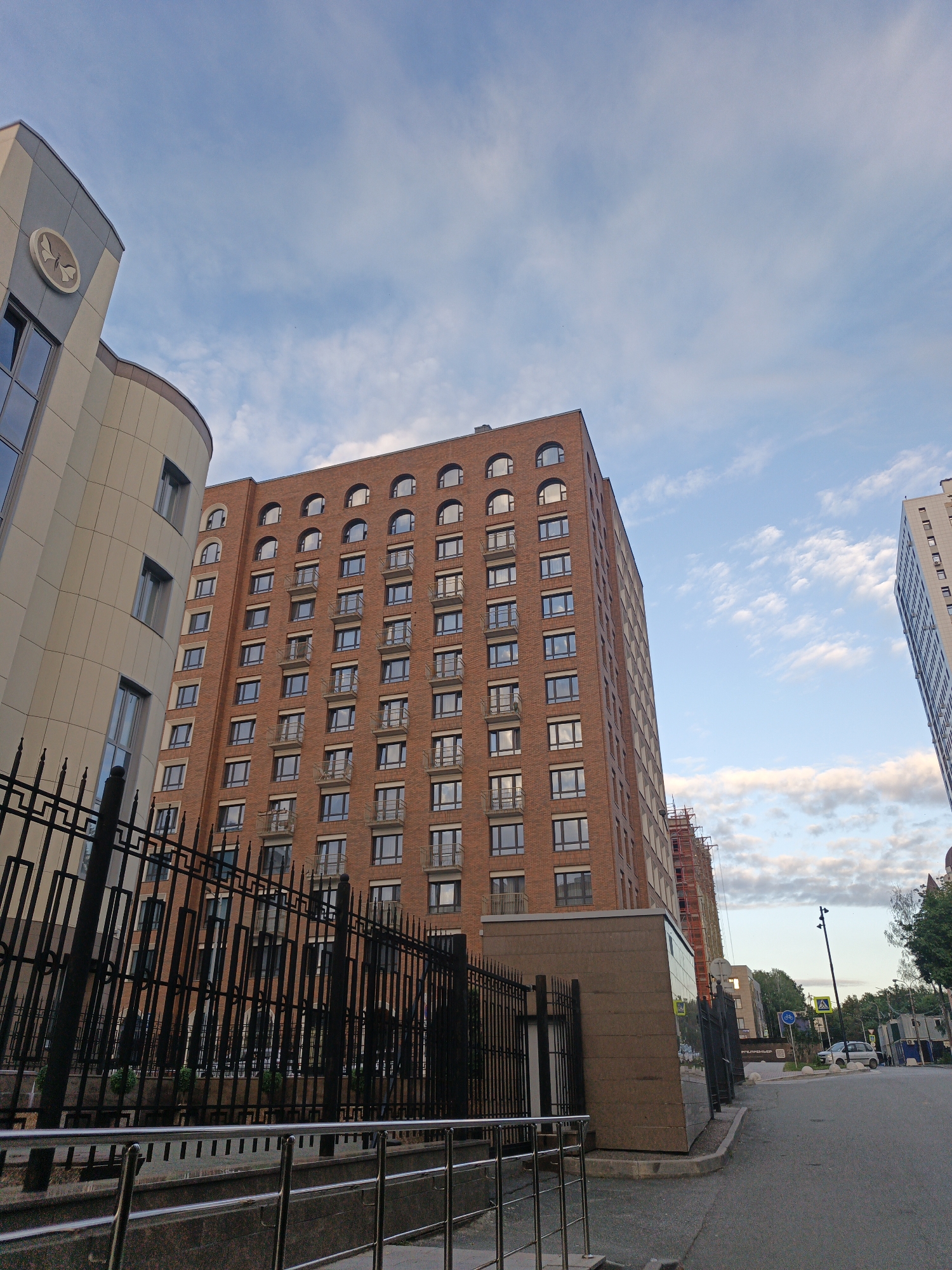 This media file captures an exterior view of several modern buildings under a cloudy sky, likely in the evening. The primary focus is on a tall, multi-story brick building with numerous windows and balconies. To its left is a curved, lighter-colored building with large windows and a circular emblem featuring a stylized butterfly or moth. Further in the background, a slimmer, white building is visible, also with many windows. A black metal fence runs along the foreground, separating the viewer from the buildings. The ground in front of the fence appears to be a paved area with some landscaping behind the fence. A street is visible to the right of the buildings, with a car parked on it and streetlights. The overall impression is one of an urban or suburban environment, possibly a residential or commercial complex. The lighting suggests it is either early morning or late afternoon, with diffused sunlight filtering through the clouds. There is no discernible activity or people in the image. No text is clearly legible.
