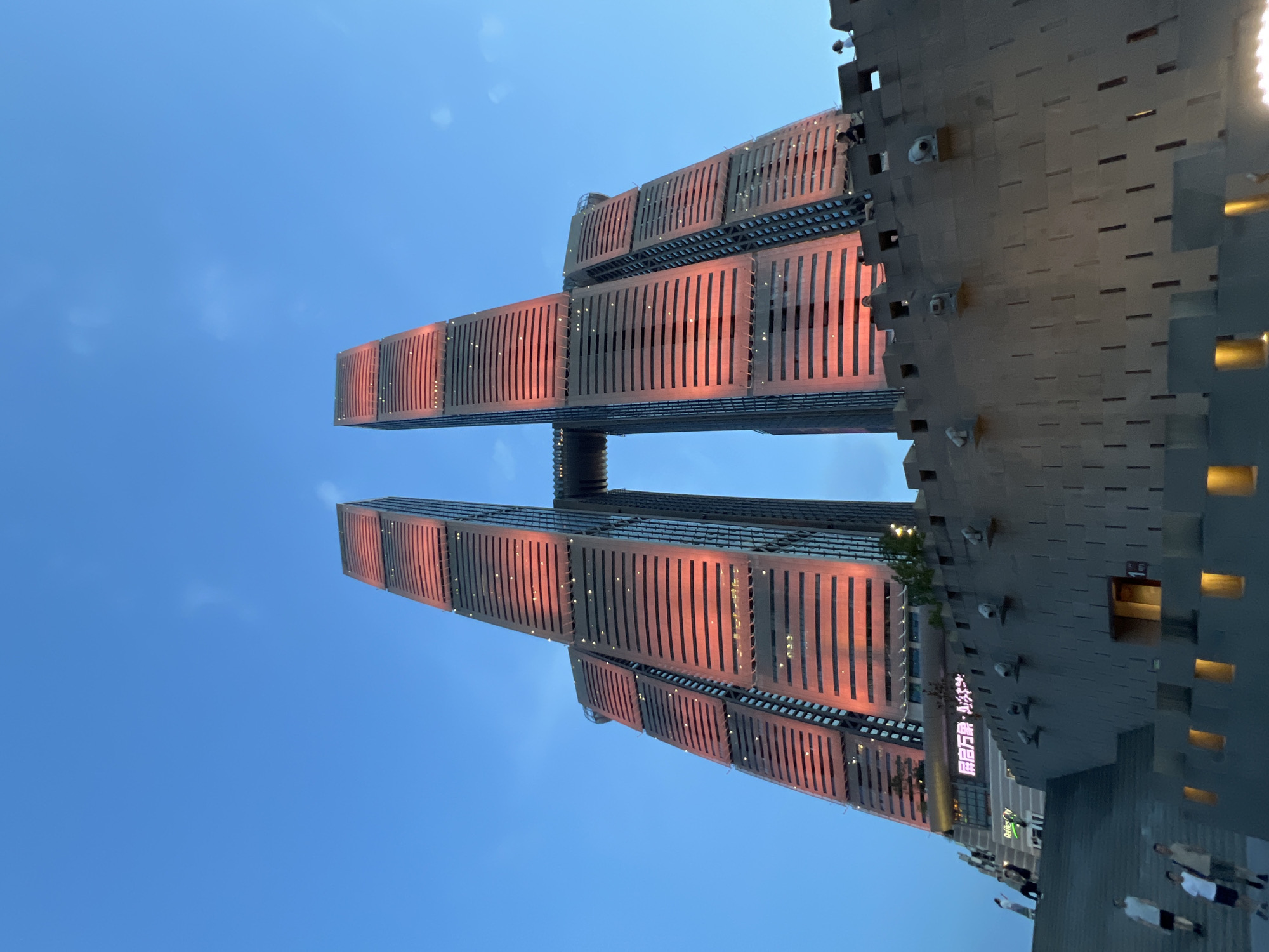 This is a low-angle, full shot of the Jinan Minhang Building in Yuzhong District, China, at dusk. The building is a modern skyscraper, distinguished by its twin towers connected by a skybridge. The facade of the towers is illuminated with a vibrant orange-red light, highlighting horizontal bands of windows. The sky is a deep blue with a hint of lighter blue near the horizon. In the foreground, a large, textured concrete wall with crenellations and decorative elements reminiscent of ancient fortifications dominates the lower part of the frame. Steps lead up to the wall on the left side of the image, where several individuals are visible walking and standing. A few people are also seen on top of the wall, observing the building. The overall scene captures the striking contrast between modern architecture and historical design elements, set against a twilight sky.
